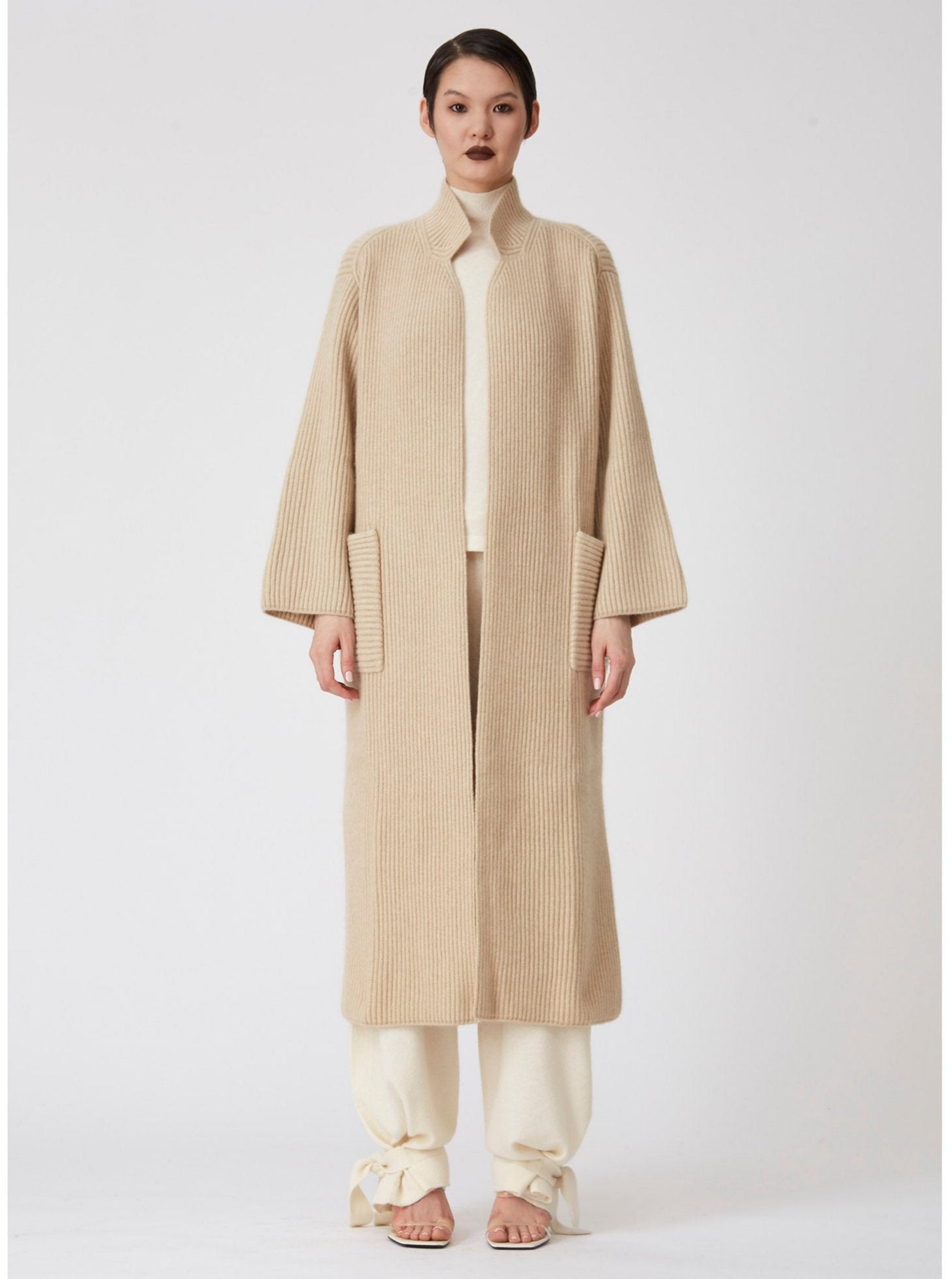 Front view of model wearing a beige Midi cardigan coat in ribbed cashmere knit. Straight cut with a relaxed fit, deep patch pockets and generous sleeves which can be rolled back.