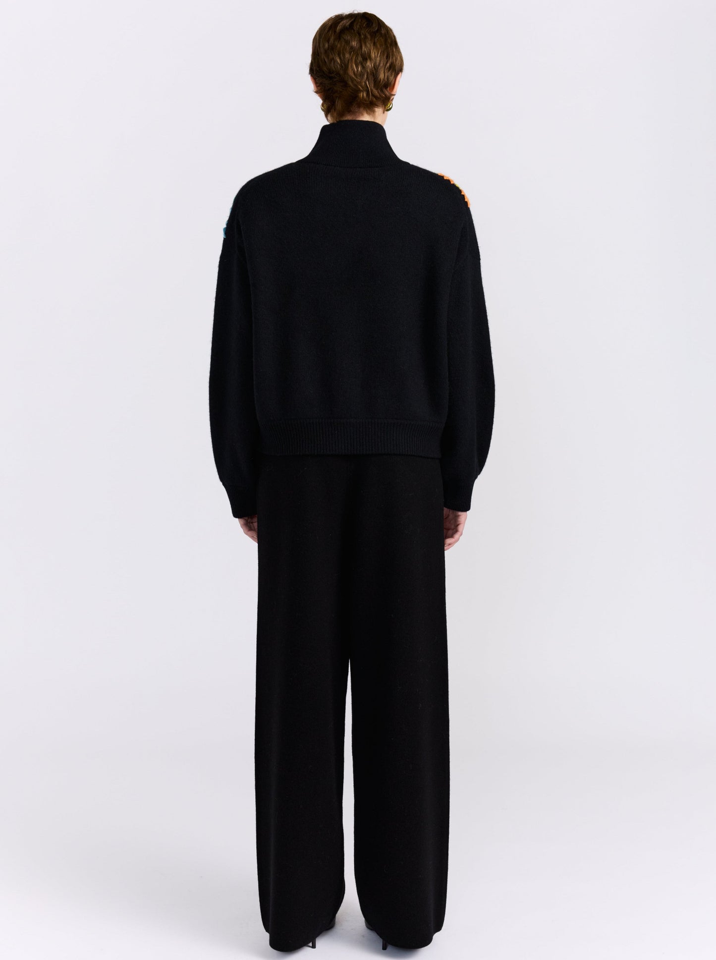 A model viewed from the back wearing a black knit bomber jacket with embroidered floral details on the shoulders and matching black wide-leg trousers, set against a plain light gray studio background.