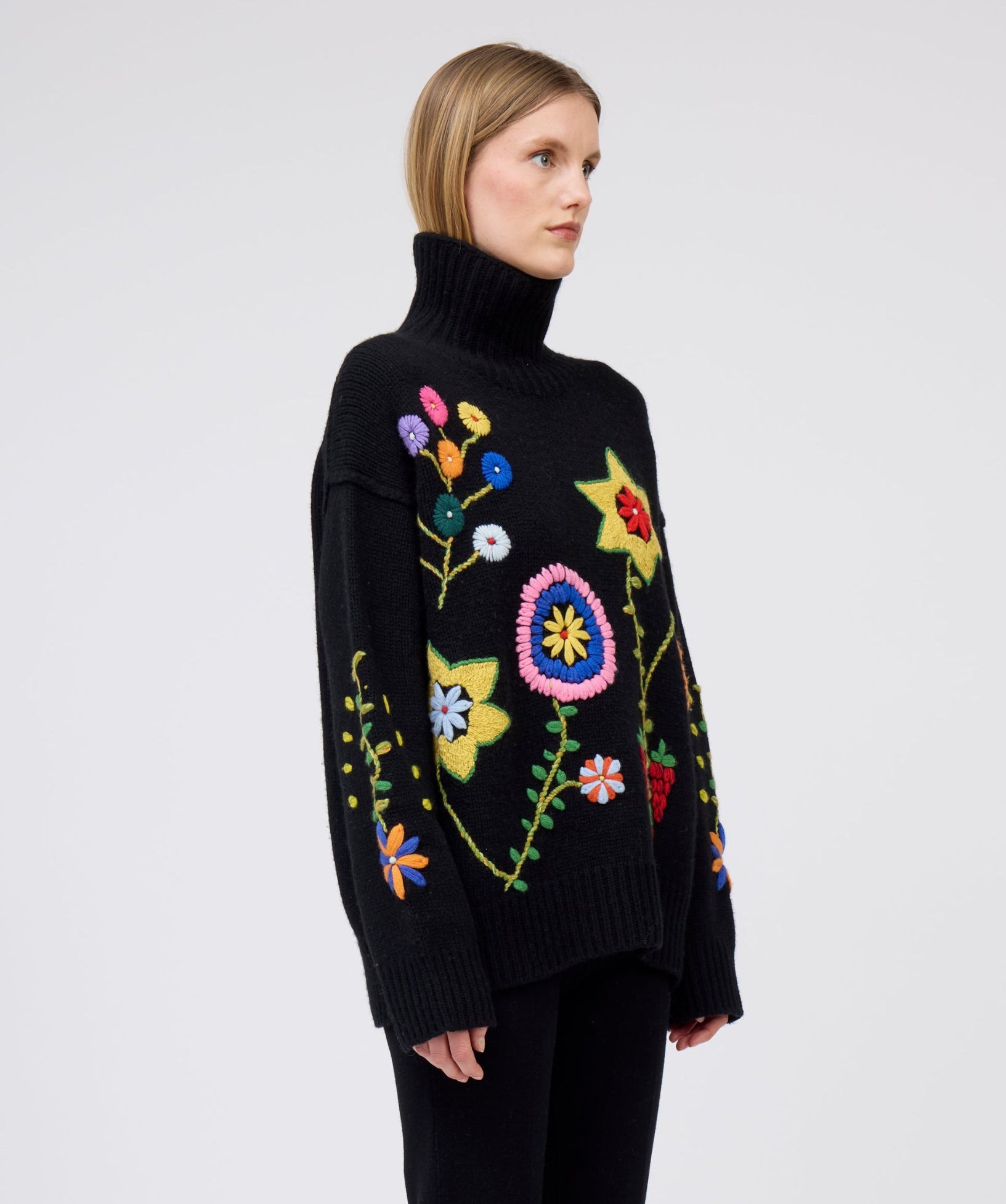 Close up of model wearing black oversized roll neck jumper adorned with brightly coloured hand embroidered flowers.