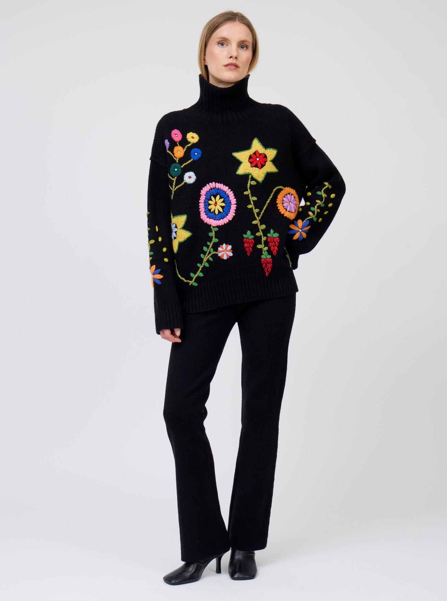 Model wearing black oversized roll neck jumper adorned with brightly coloured hand embroidered flowers; worn with black cashmere suit pants.