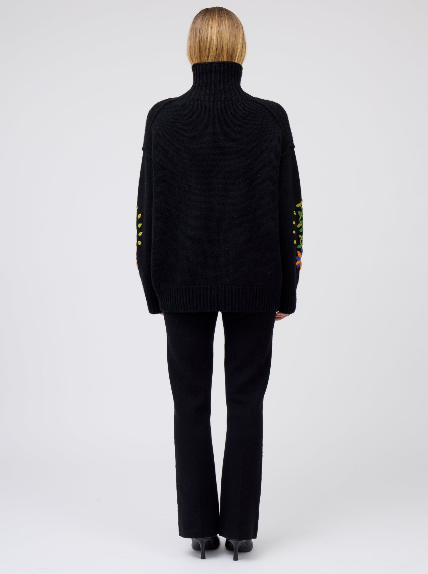 Back view of model wearing black oversized roll neck cashmere jumper with some embroidery visible on the sleeves; worn with black pants.