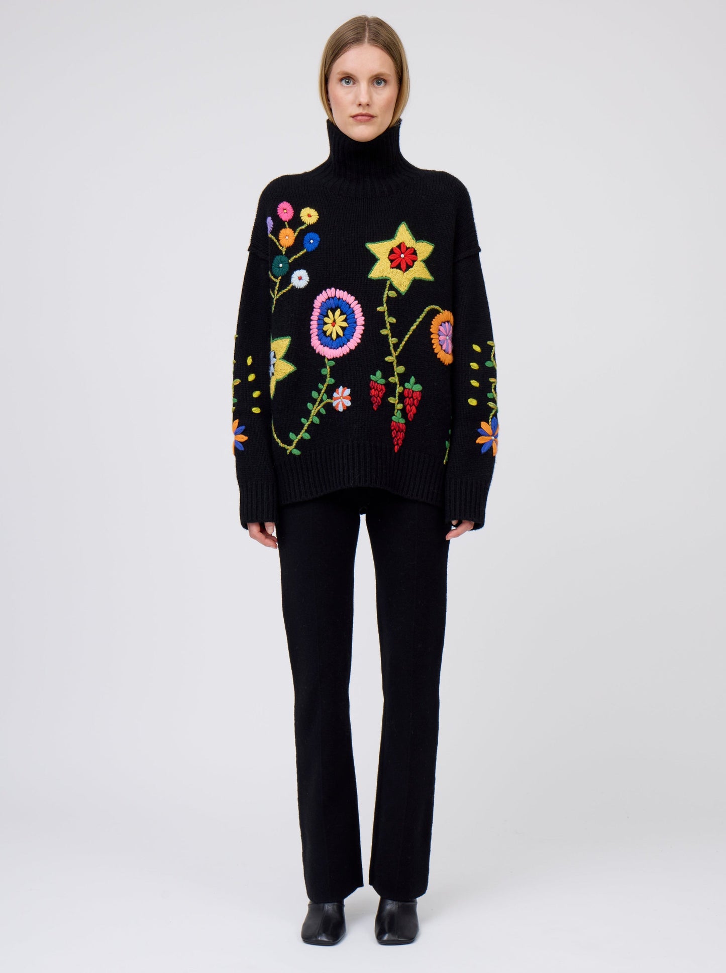Front view of model wearing black oversized roll neck jumper adorned with brightly coloured hand embroidered flowers.