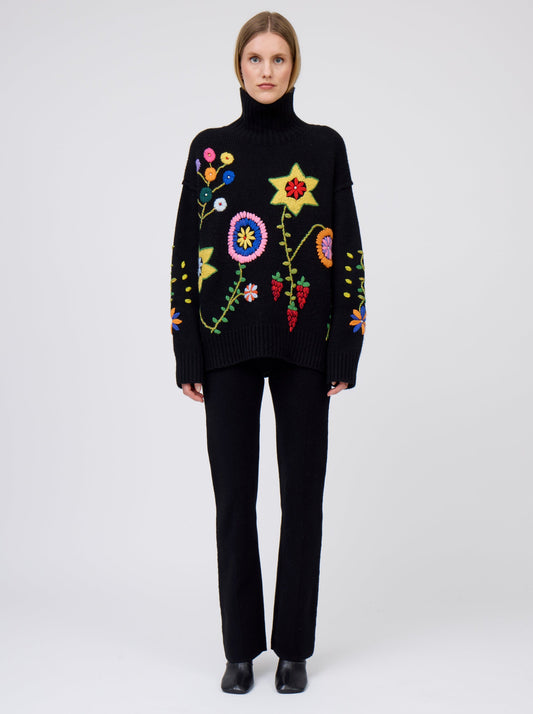 Front view of model wearing black oversized roll neck jumper adorned with brightly coloured hand embroidered flowers.