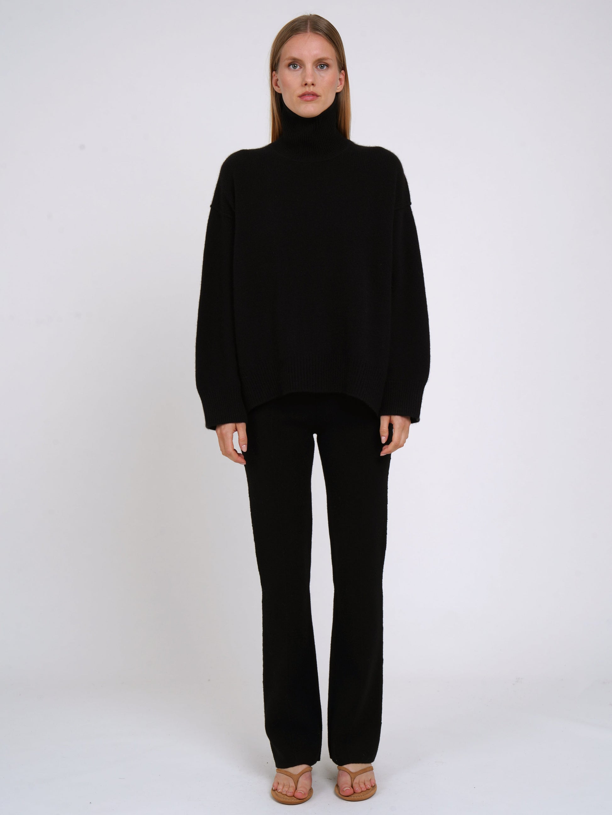 Front view of model wearing black cashmere oversized roll neck jumper with generous long sleeves and high-low hem; worn with black cashmere tailored pants.
