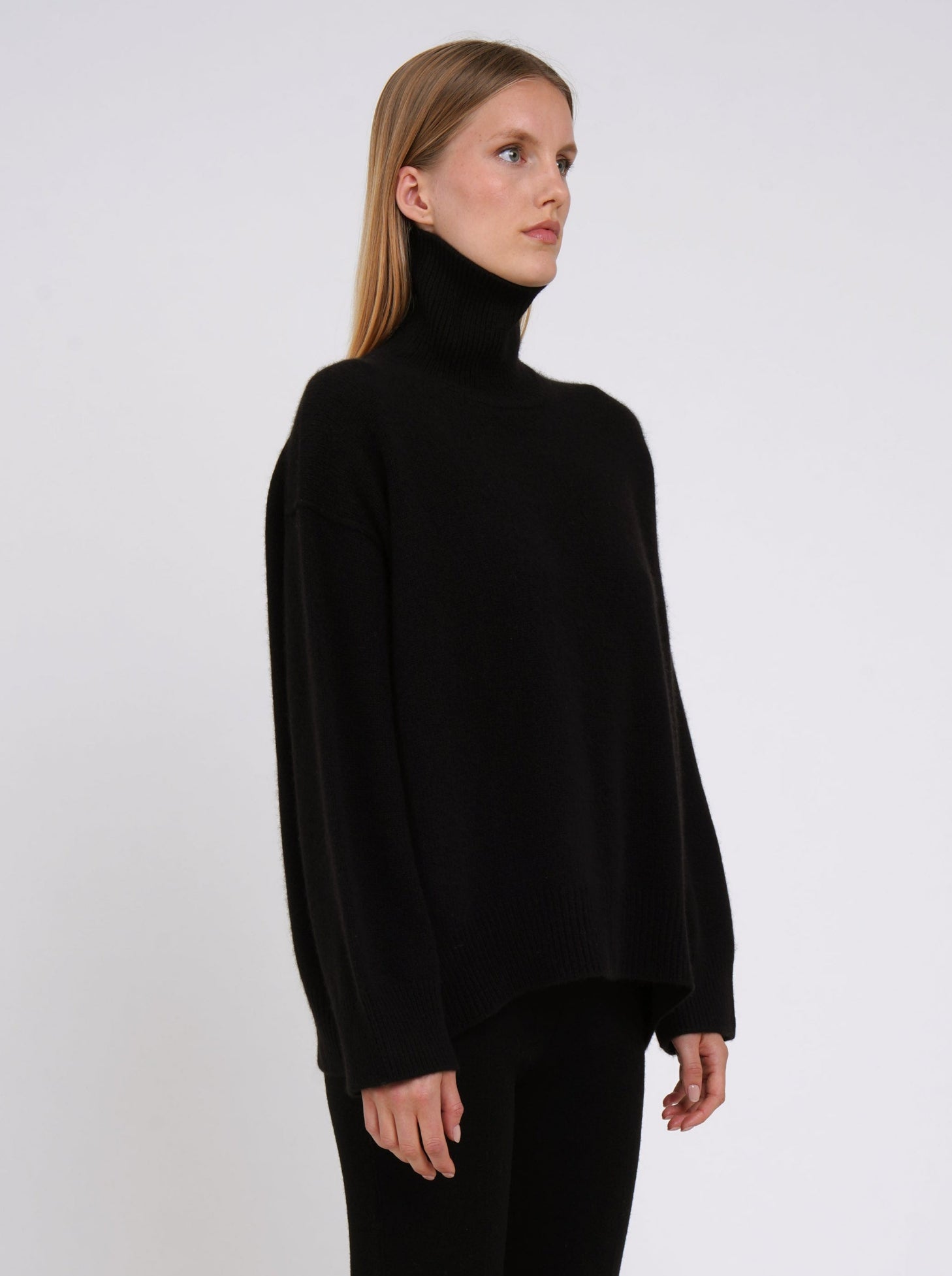 Close-up of model wearing black cashmere oversized roll neck jumper with generous long sleeves and high-low hem.
