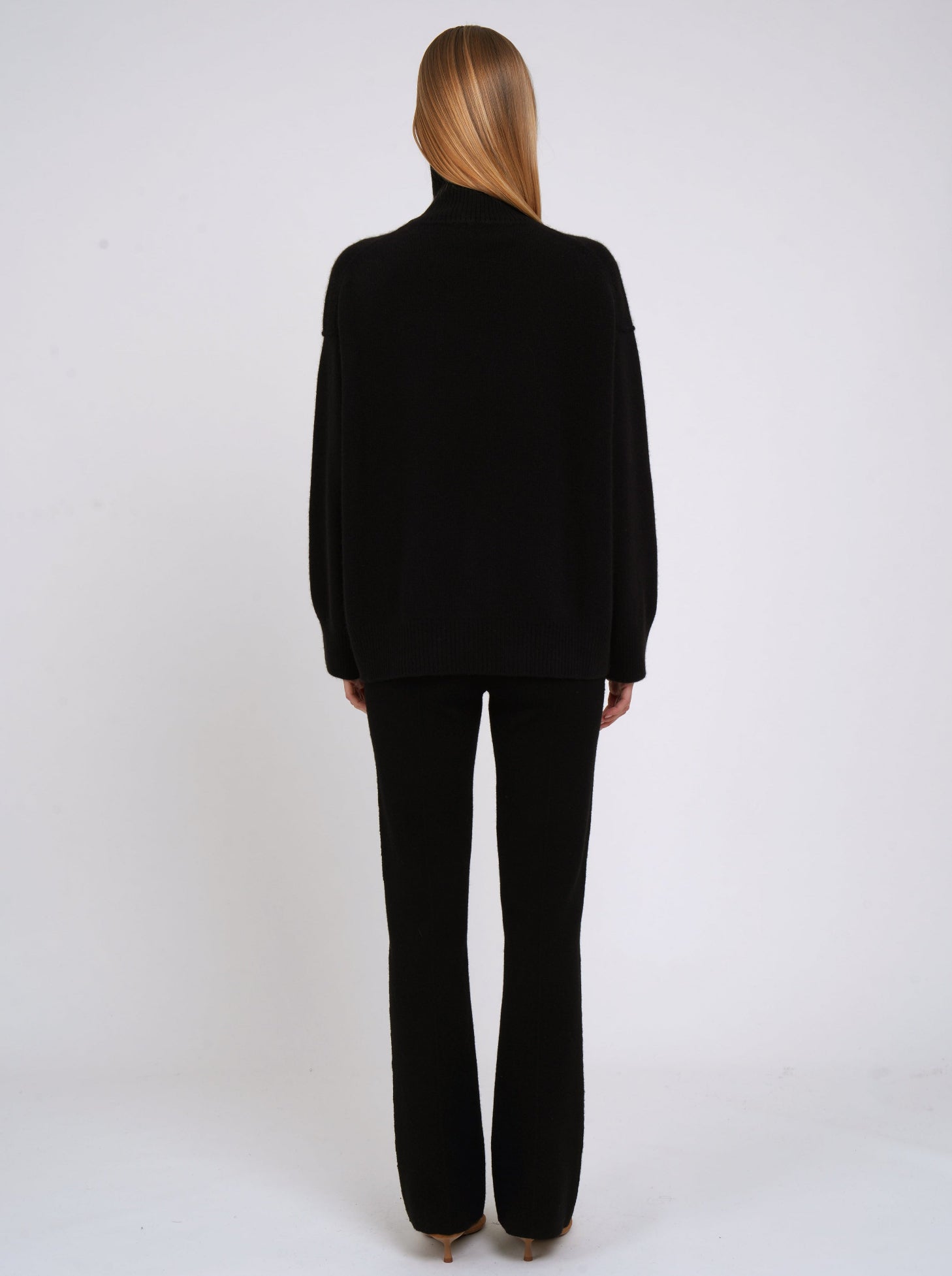 Back view of model wearing black cashmere oversized roll neck jumper with generous long sleeves and high-low hem; worn with black cashmere tailored pants.