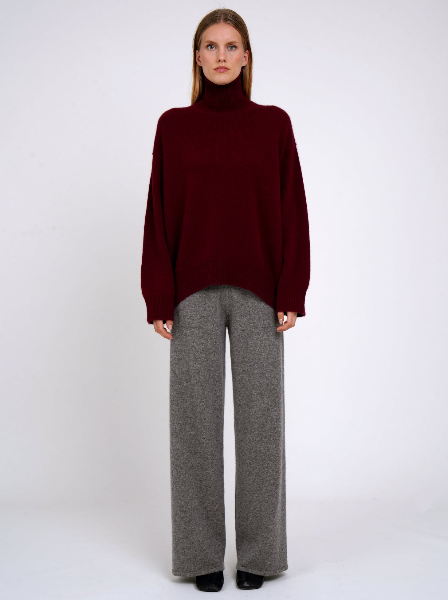 Front view of model wearing burgundy cashmere oversized roll neck jumper with generous long sleeves and high-low hem; worn with grey cashmere wide leg pants.