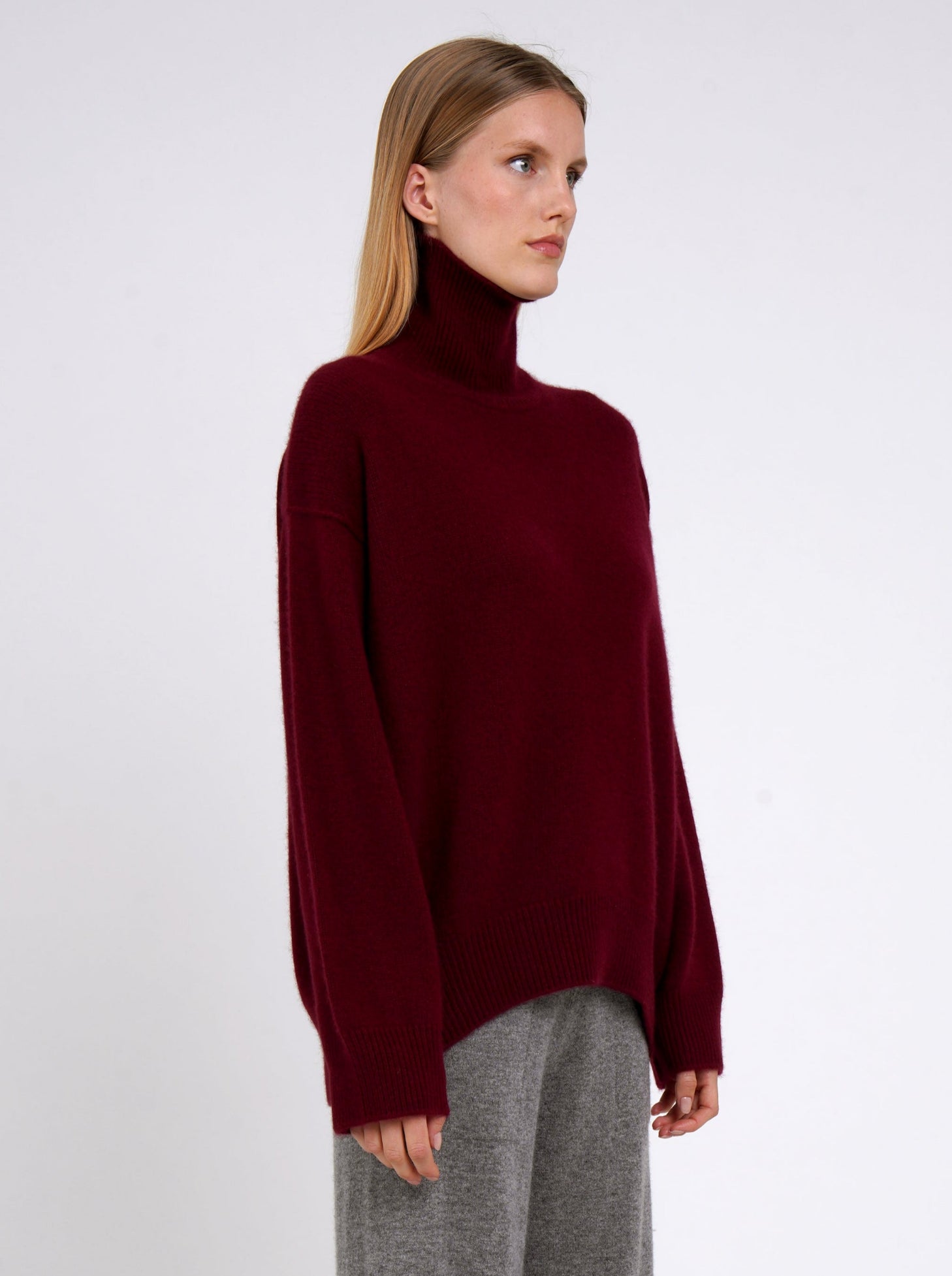 Close up model wearing burgundy cashmere oversized roll neck jumper; worn with grey cashmere wide leg pants.