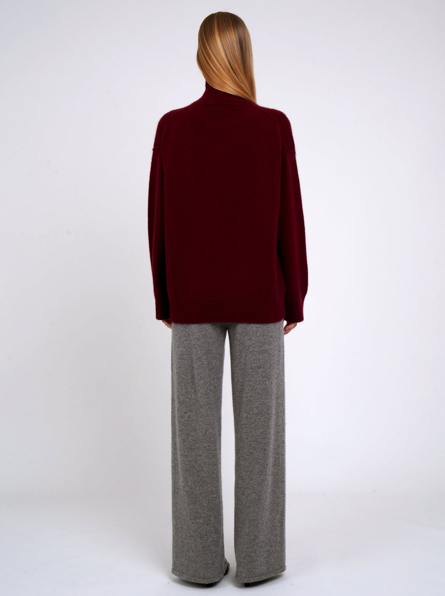 Back view of model wearing burgundy cashmere roll neck oversized jumper; worn with grey cashmere wide leg pants.