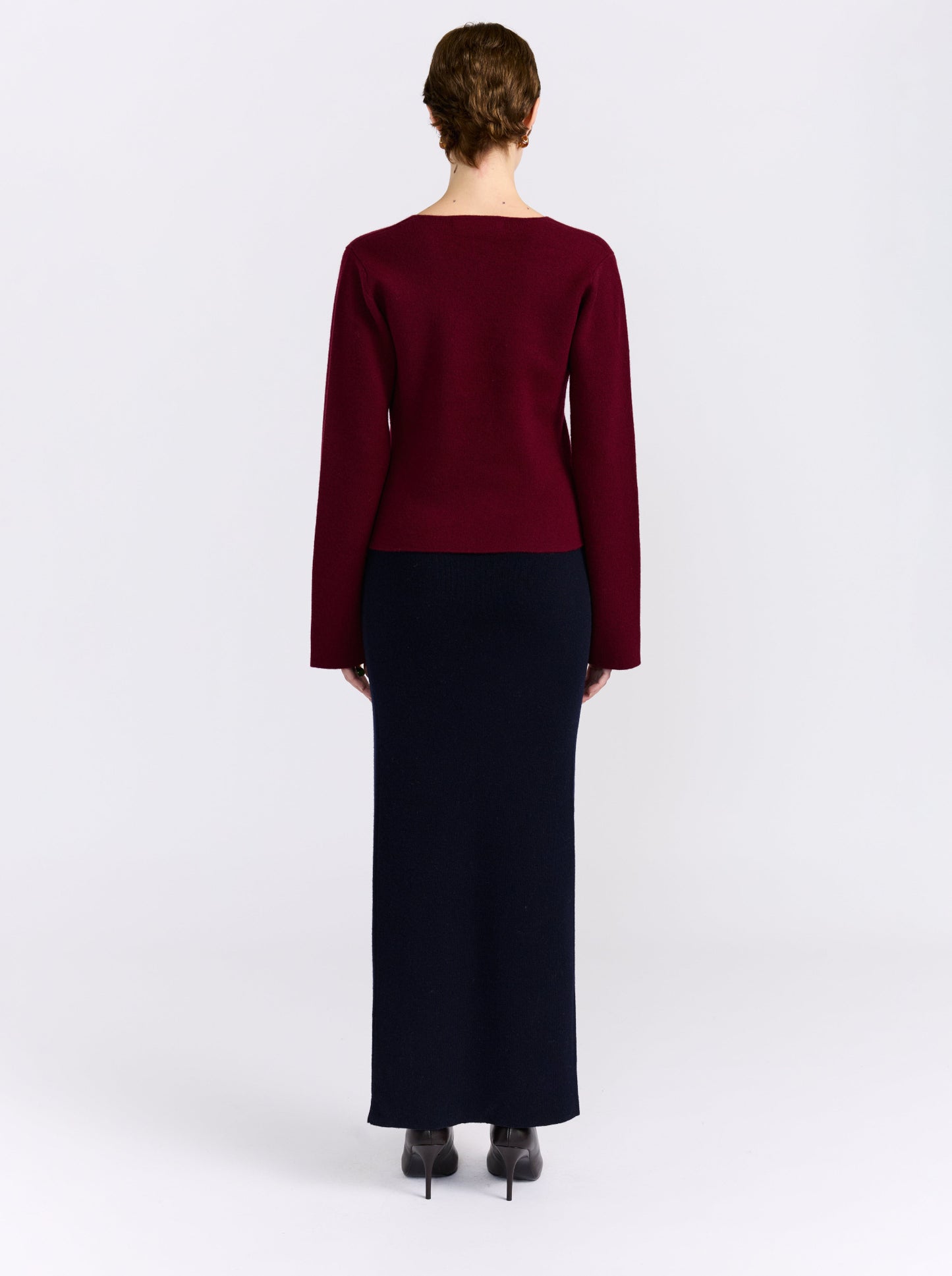 A model shown from the back wearing a long-sleeve knit top in deep burgundy and a long dark navy skirt, paired with black heels, against a plain light gray studio background.