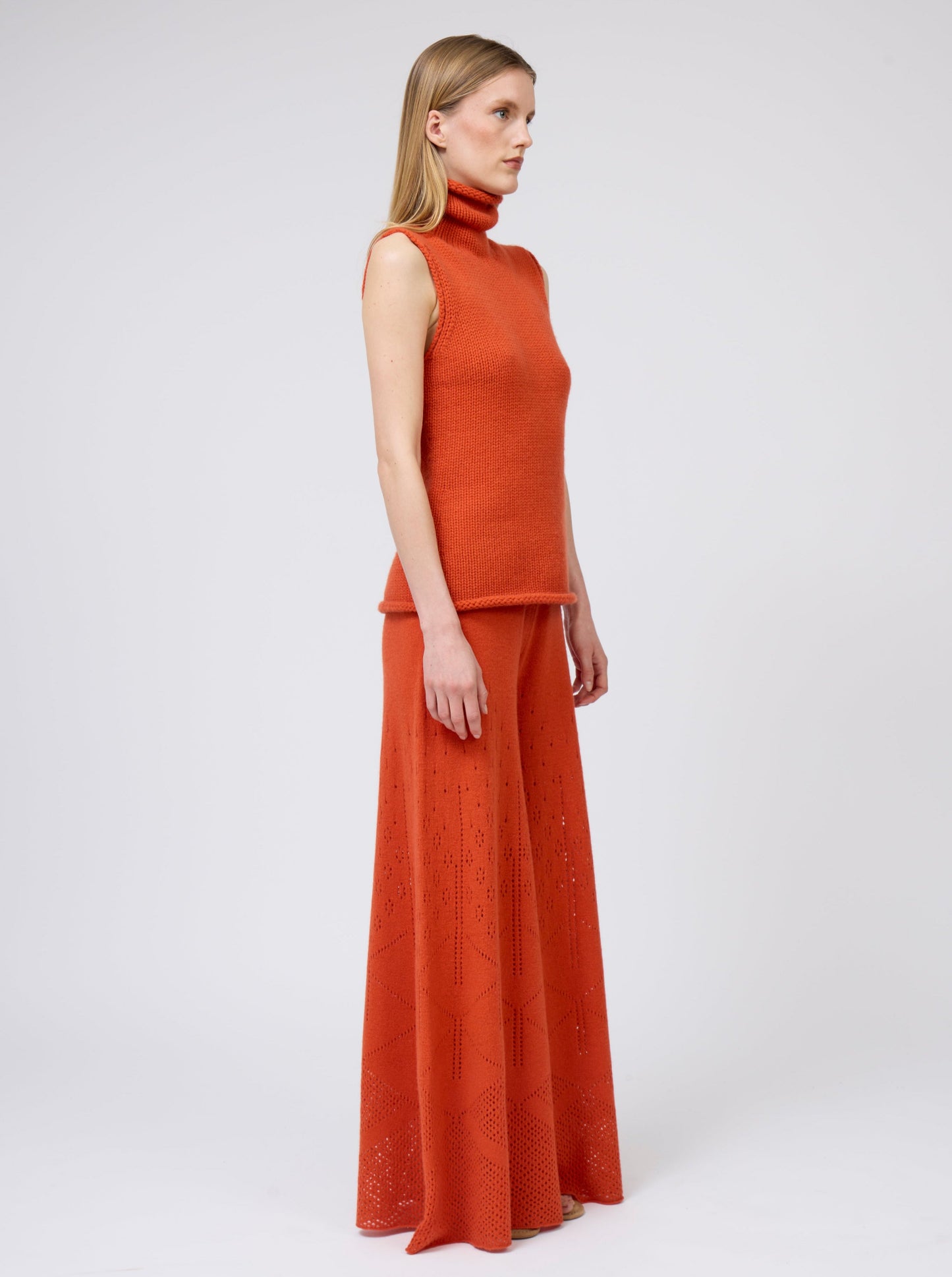 Side view model in orange cashmere chunky knit sleeveless turtle neck tank top with matching orange full flared trousers.