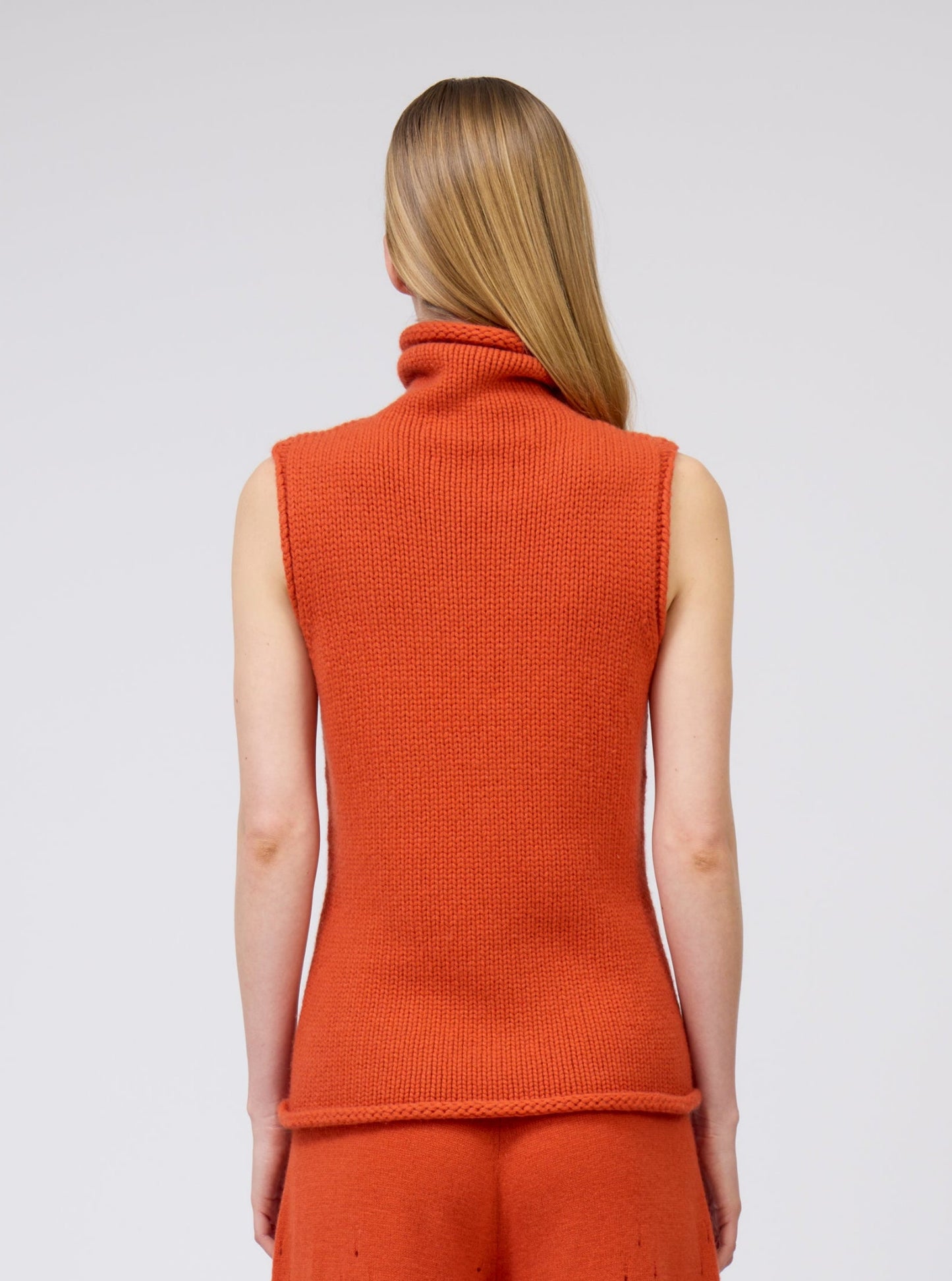 Cropped back view model in orange cashmere chunky knit sleeveless turtle neck tank top with matching orange knit pants.