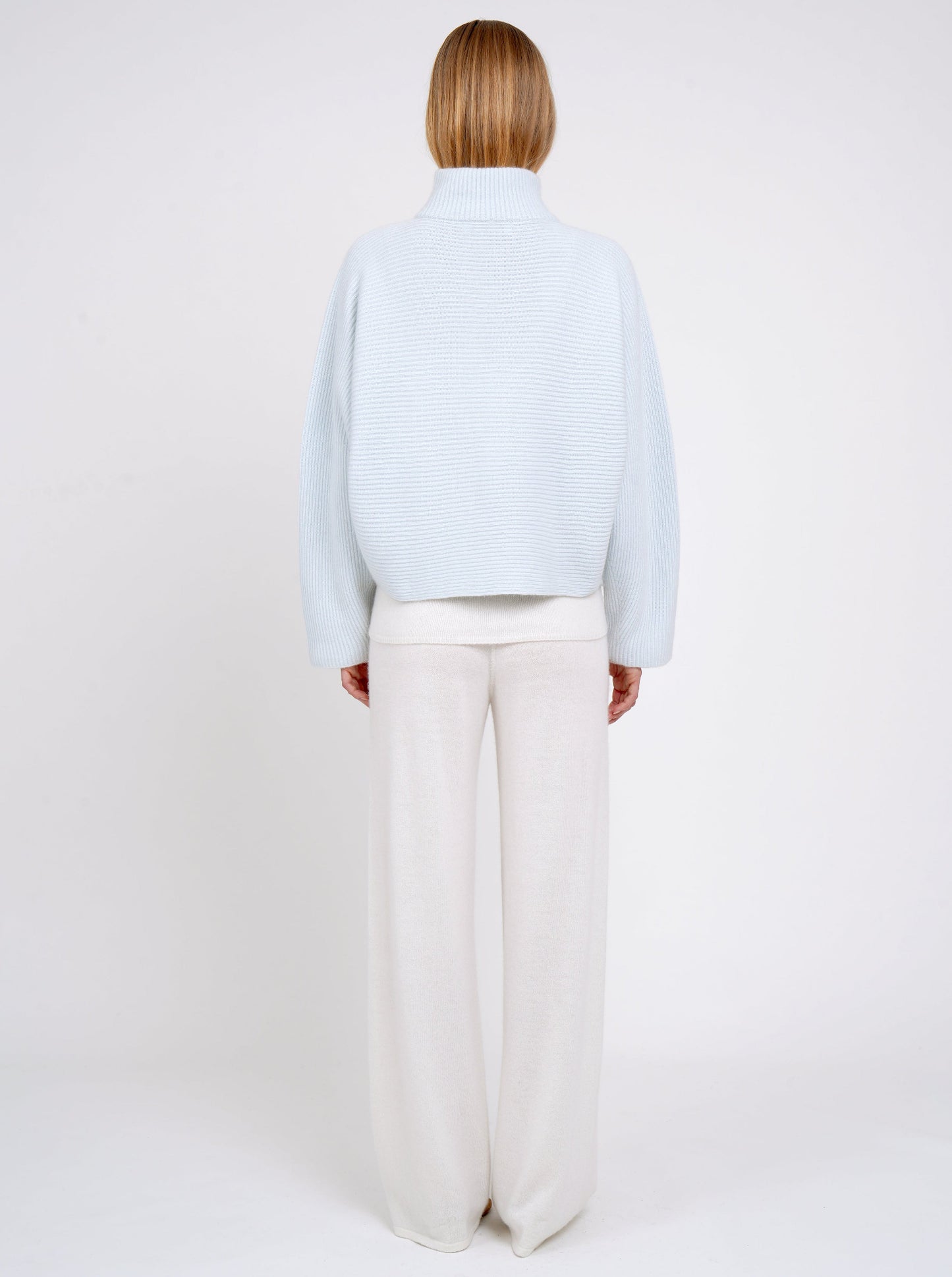 Back view model in pale blue ribbed knit cashmere high collar jacket with wide kimono sleeves; worn with white cashmere wide leg pants.