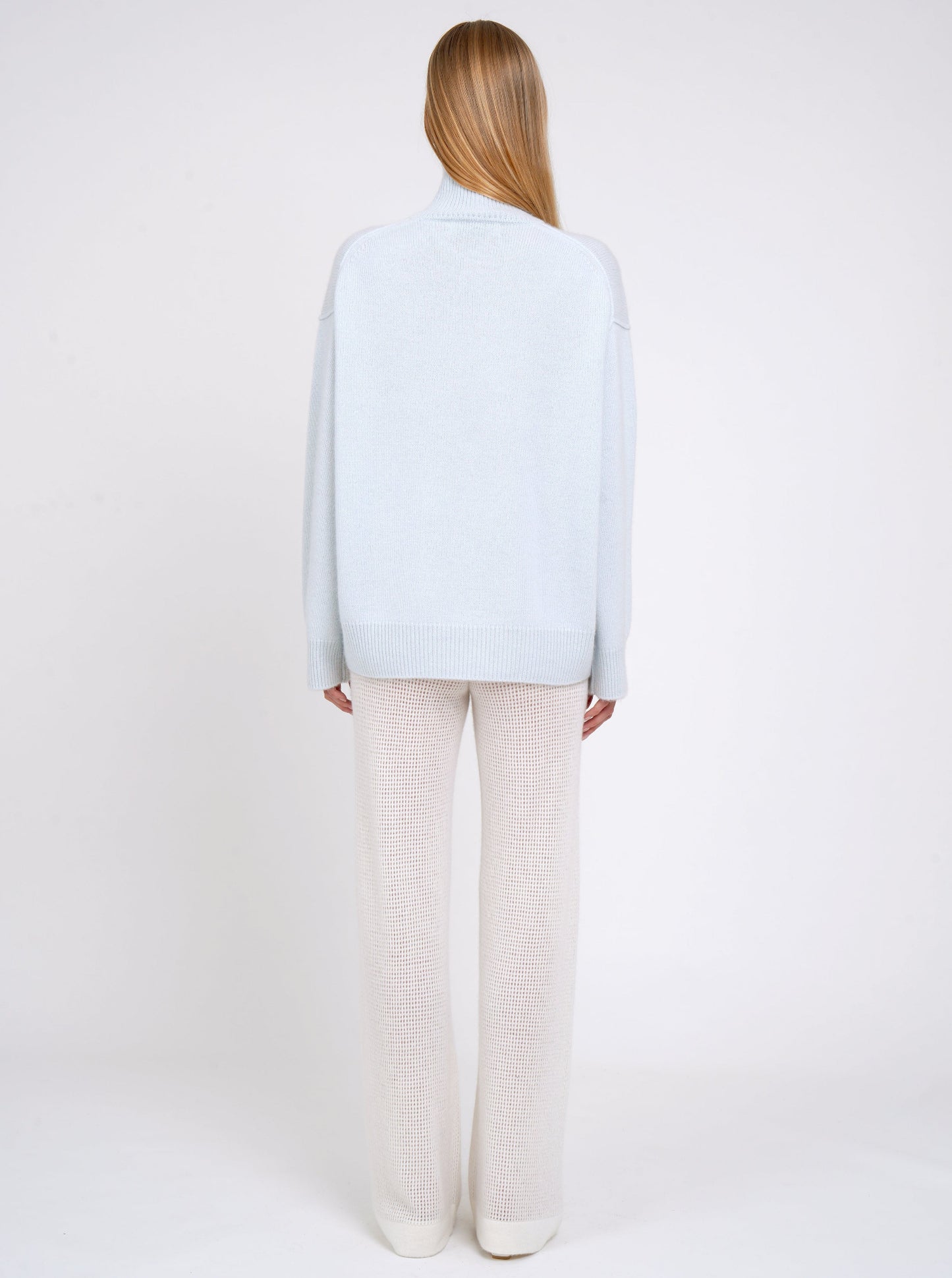 Back view of model in pale blue cashmere oversized roll neck jumper with generous long sleeves and high-low hem; worn with white cashmere, net knit, wide leg pants.