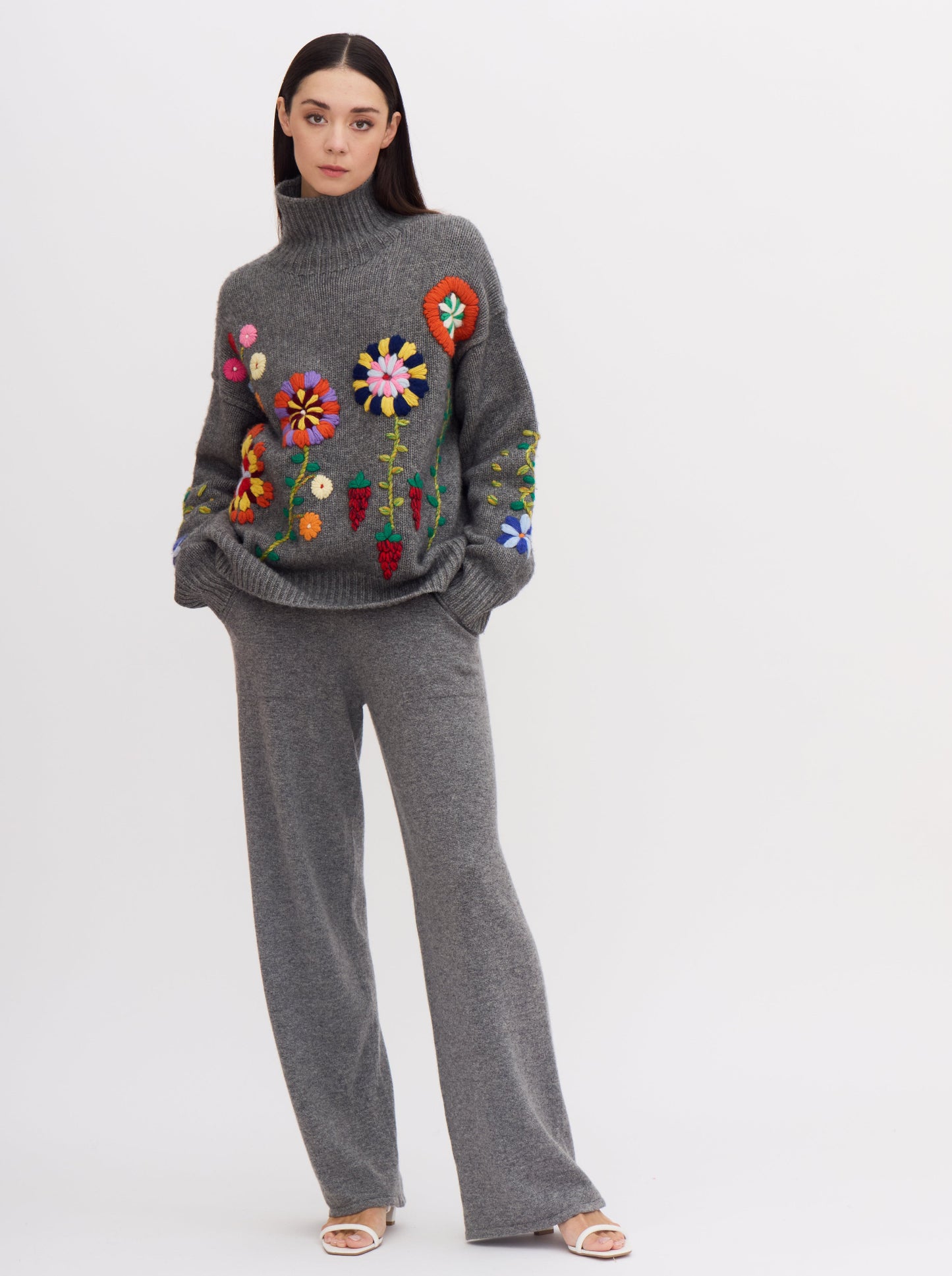 Model wears relaxed, grey cashmere, wide leg pants with pockets; worn with grey cashmere chunky knit oversize jumper with bright colour hand embroidered flowers.
