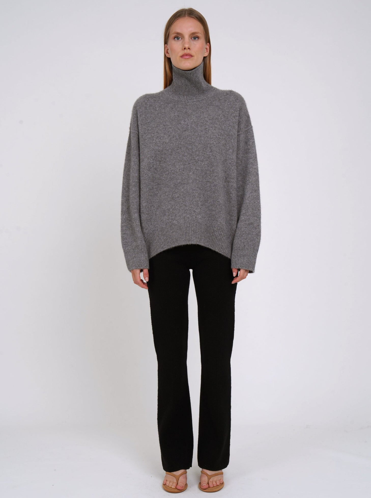 Front view of model wearing grey cashmere oversized roll neck jumper with generous long sleeves and high-low hem; worn with black cashmere tailored pants.
