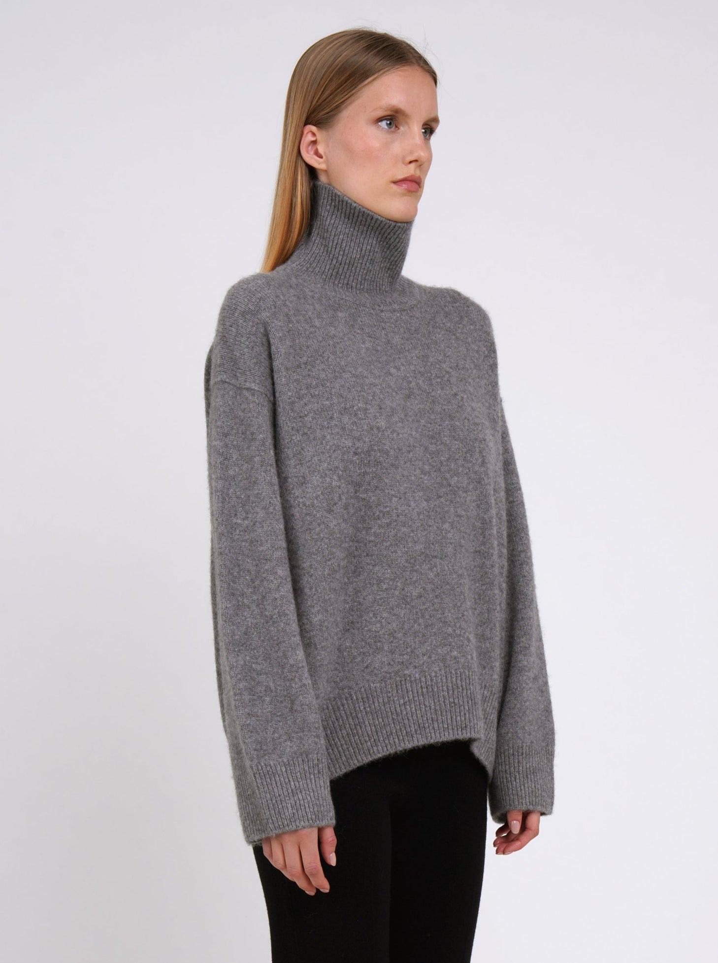 Close-up of model wearing grey cashmere oversized roll neck jumper with generous long sleeves and high-low hem.