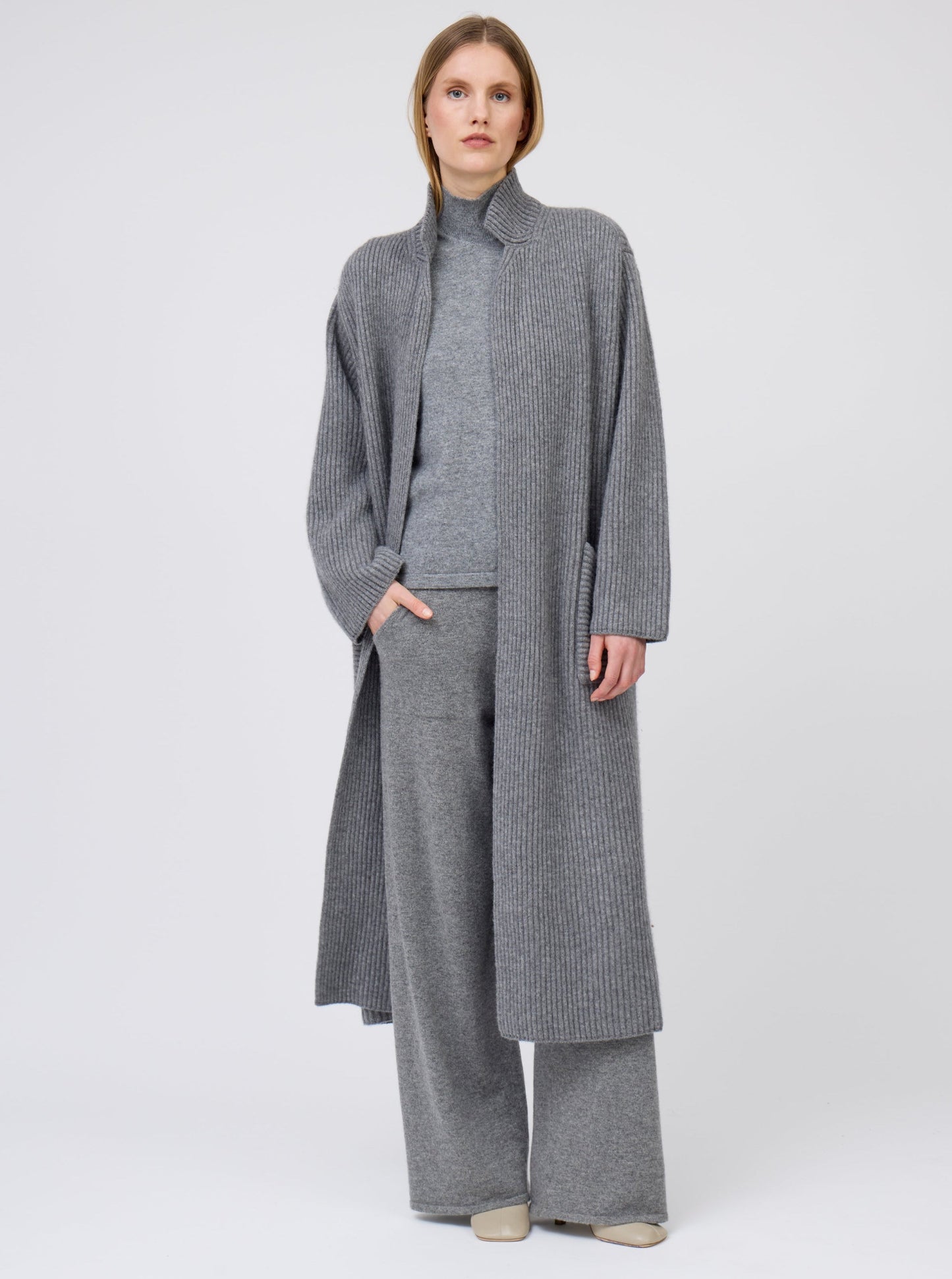 Model wearing a long grey cardigan coat in ribbed knit cashmere. Straight cut with a relaxed fit, deep patch pockets; standing with her hand in one pocket of matching grey cashmere pants.