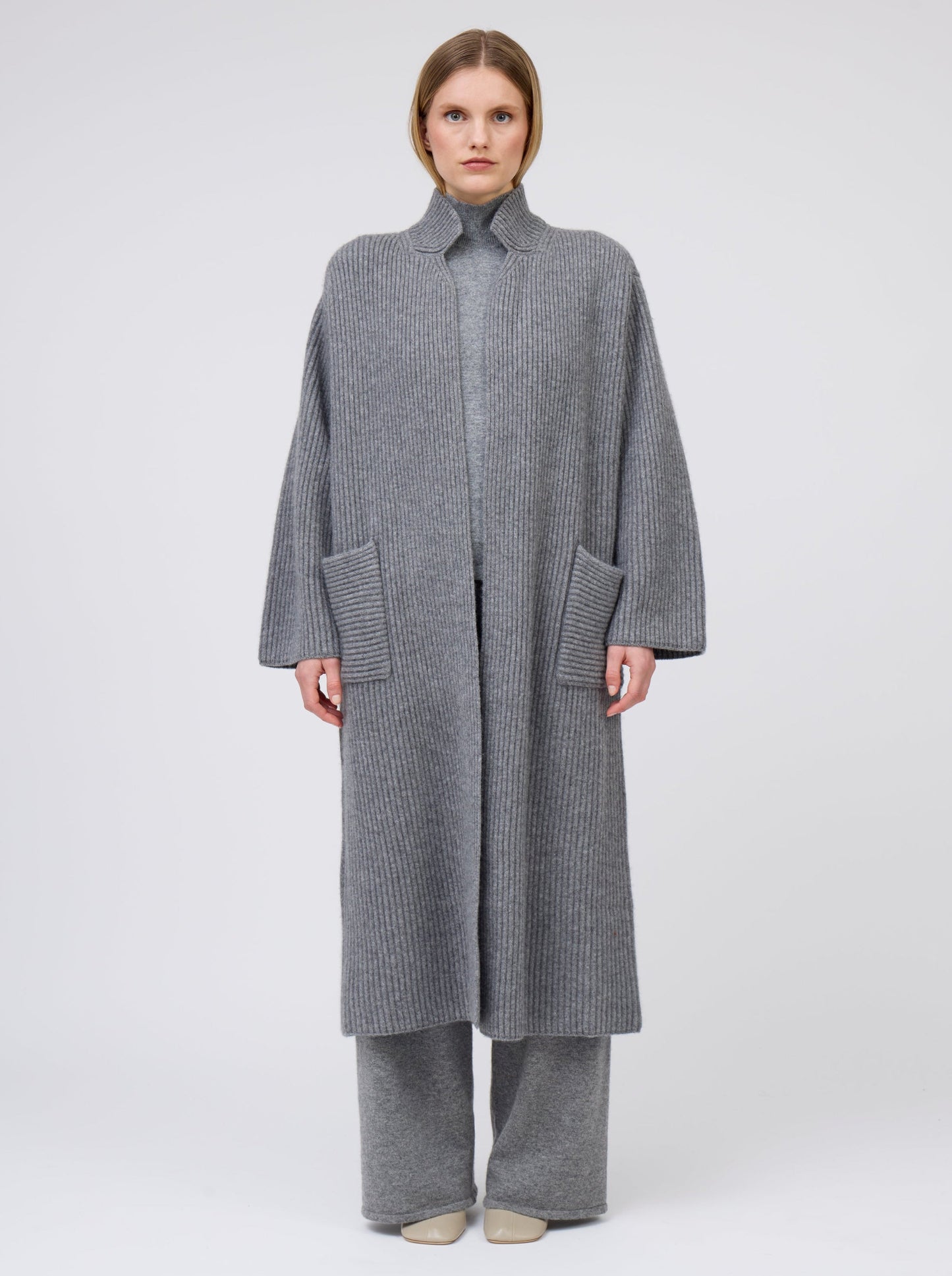 Front view of model wearing a long grey cardigan coat in ribbed knit cashmere. Straight cut with a relaxed fit, deep patch pockets and generous long sleeves. Worn with wide leg grey cashmere pants.