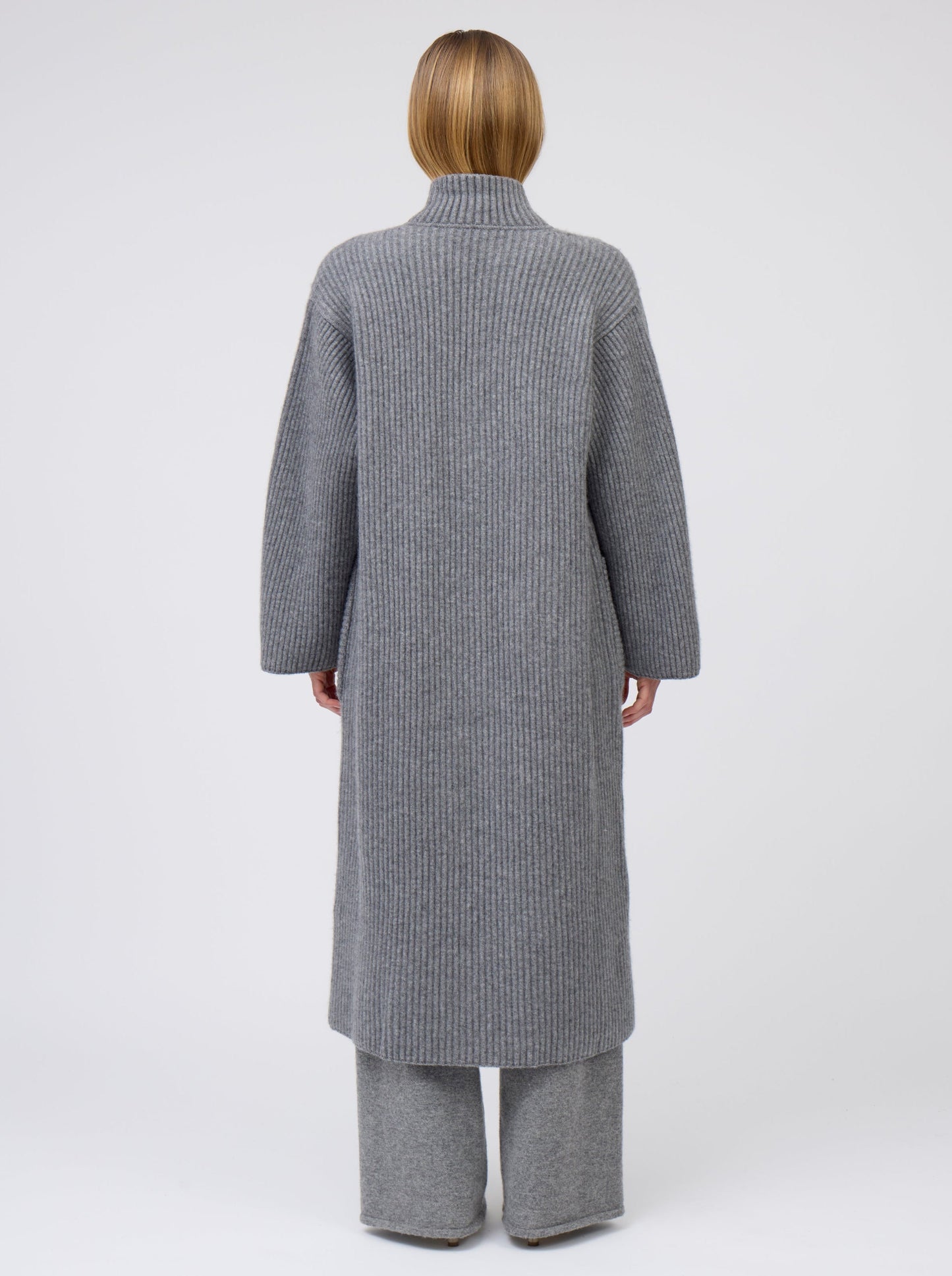 Back view of model wearing a long grey cardigan coat in ribbed knit cashmere. Straight cut with a relaxed fit, deep patch pockets and worn with collar turned up.