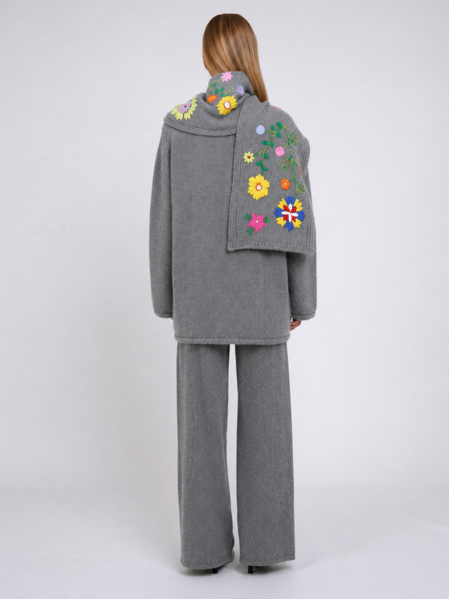 A model viewed from the back wearing a grey cashmere jacket with colourful hand embroidered florals,, paired with matching grey wide-leg trousers, against a light grey studio background.