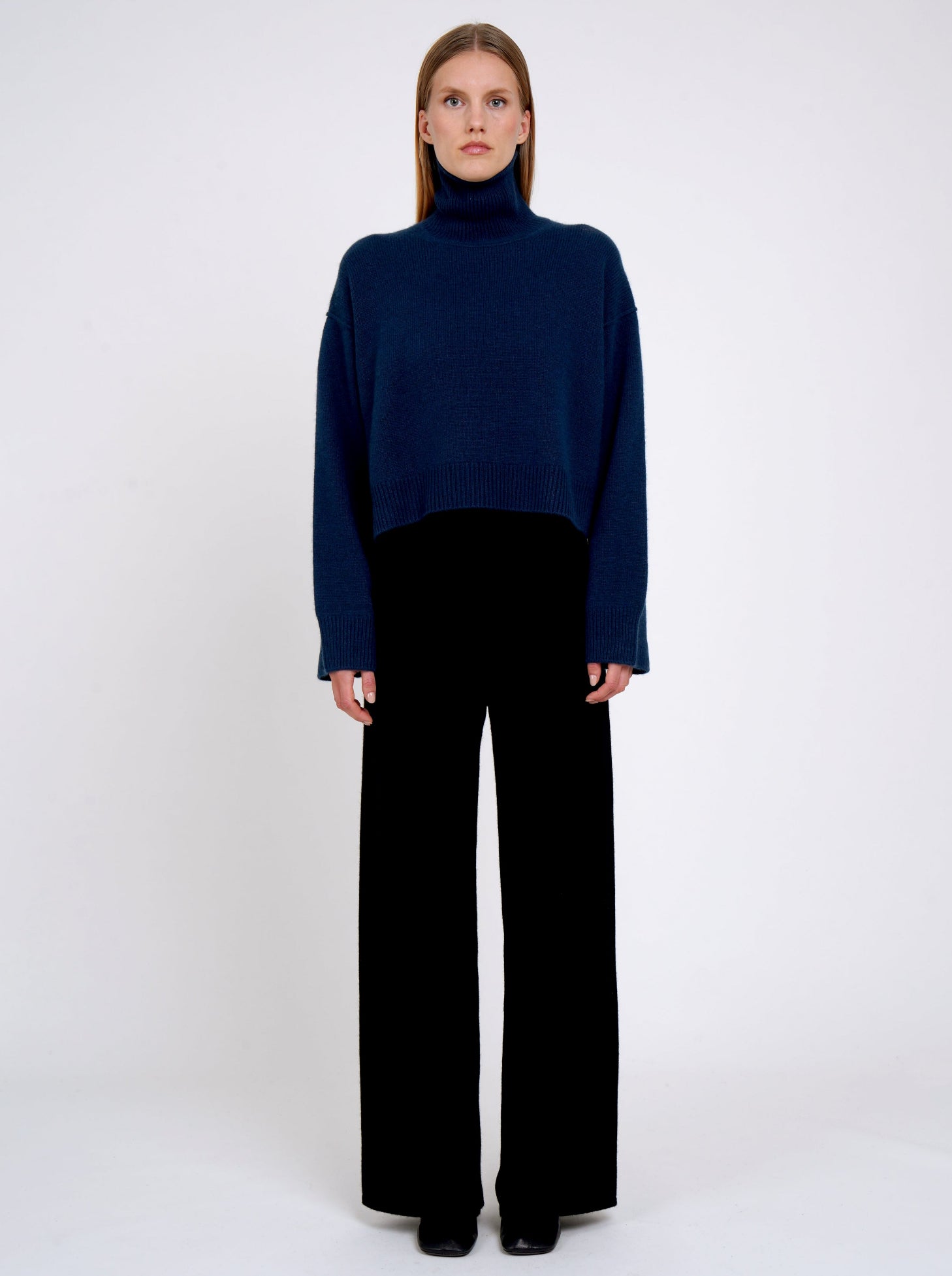 Front view of model wearing jean navy cashmere cropped length roll neck jumper with generous long sleeves and high-low hem; worn with  black cashmere wide leg pants.