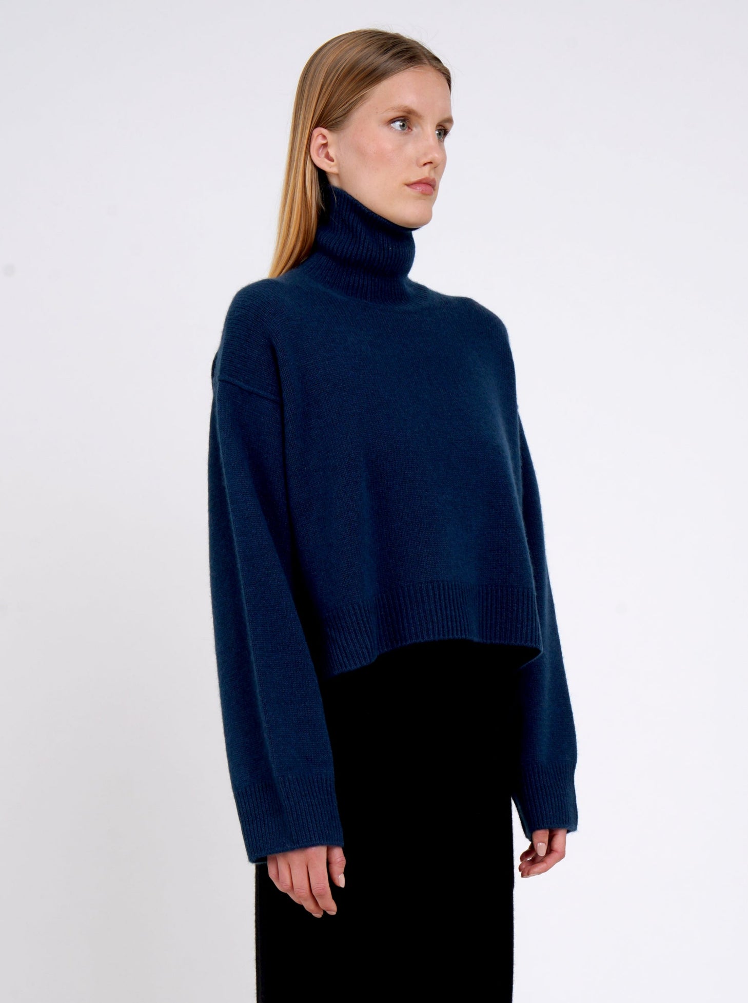 Close up of model wearing jean navy cashmere cropped length roll neck jumper with generous long sleeves and high-low hem.