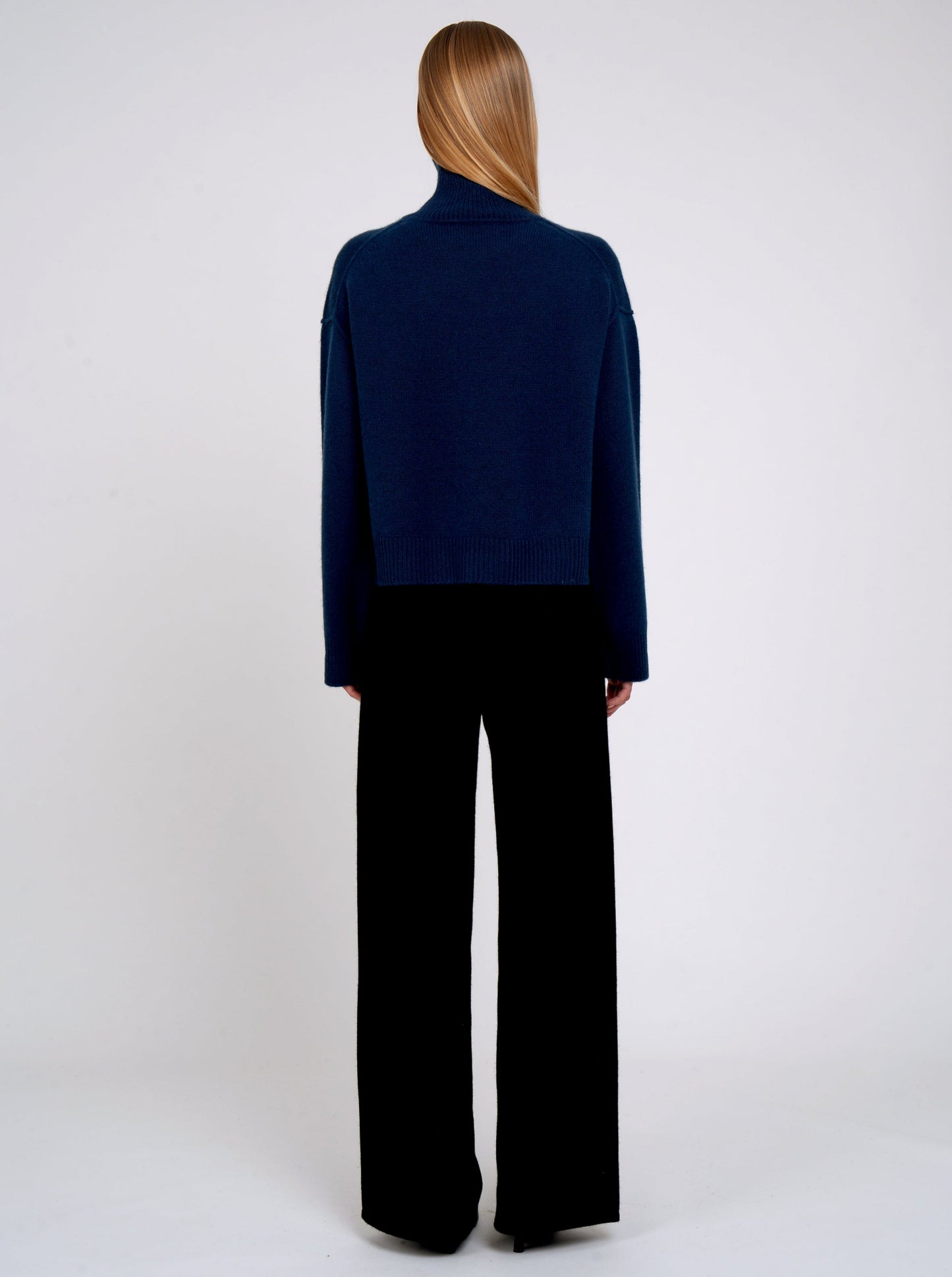Back view of model wearing jean navy cashmere cropped length roll neck jumper with generous long sleeves; worn with black cashmere wide leg pants.