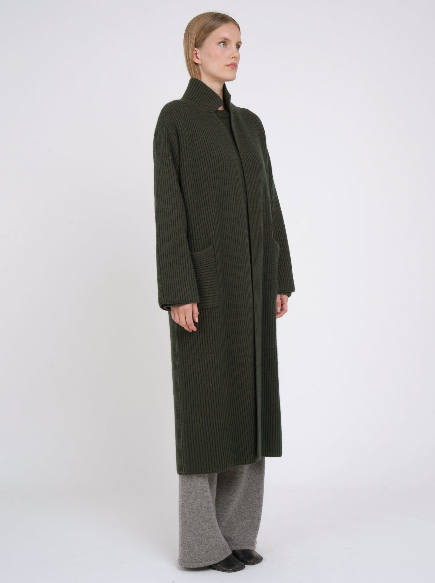 Side view model wearing a long khaki green cardigan coat in ribbed knit cashmere with high collar, patch pockets and generous long sleeves; worn with grey cashmere pants.