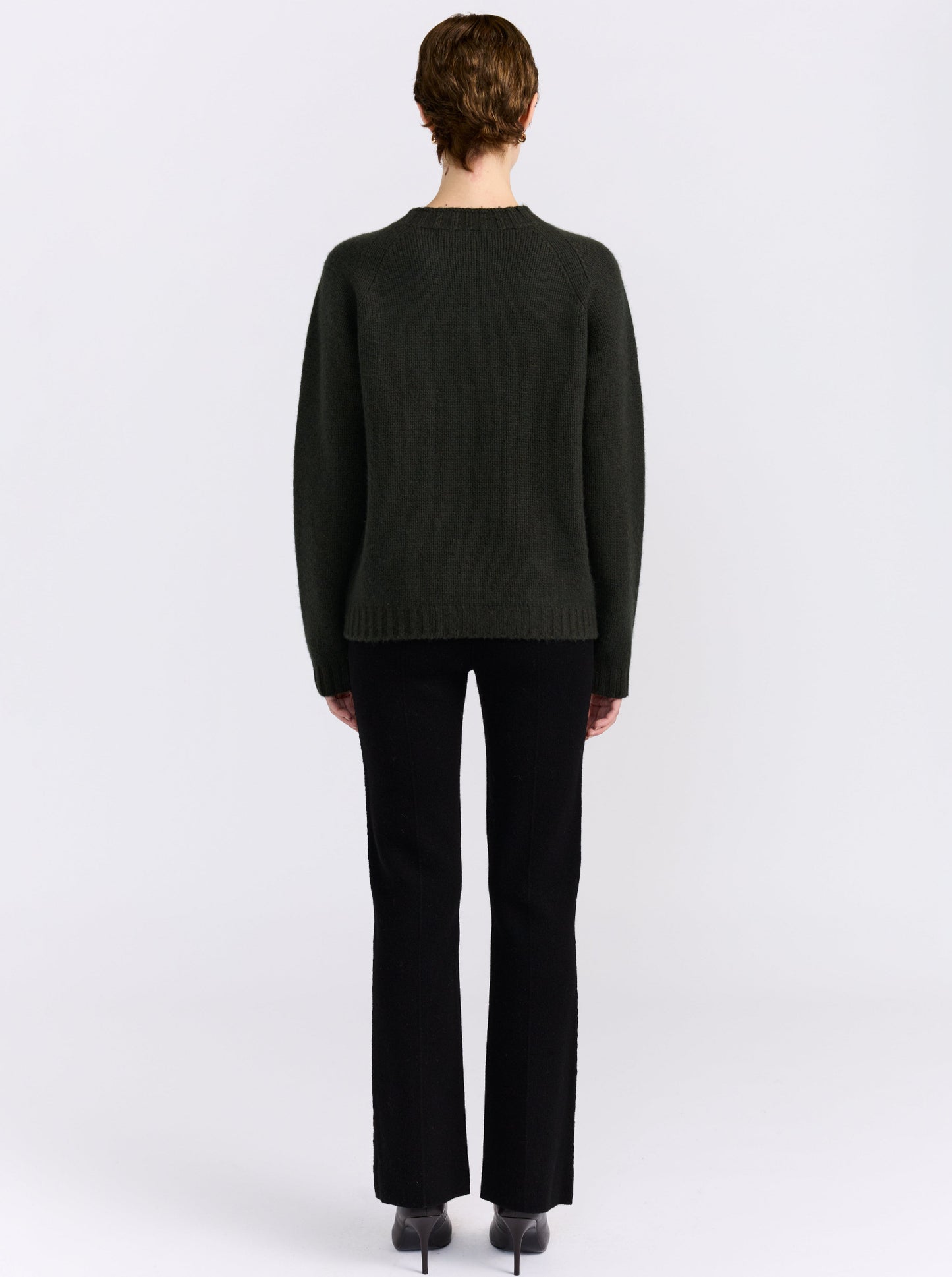 A model stands against a light gray studio background wearing khaki green cashmere round neck sweater, black pants, and black shoes.