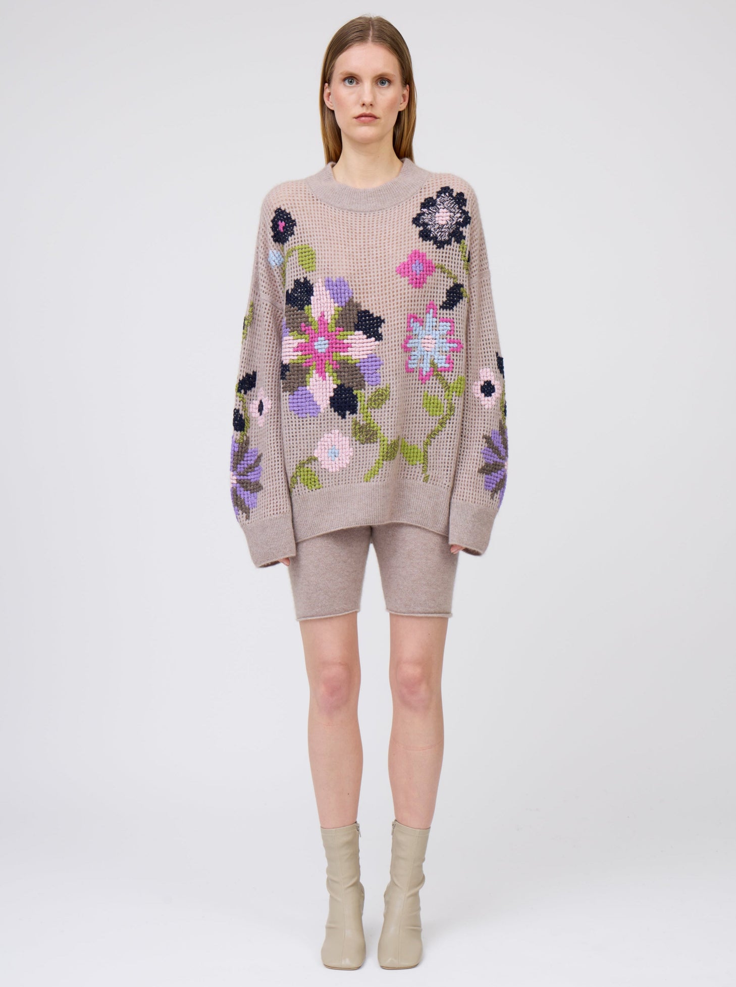 Font view of model wearing light weight beige cashmere net knit jumper, with round neck, long sleeves and bold cross-stitch floral hand embroidery; worn with beige cashmere biker shorts.
