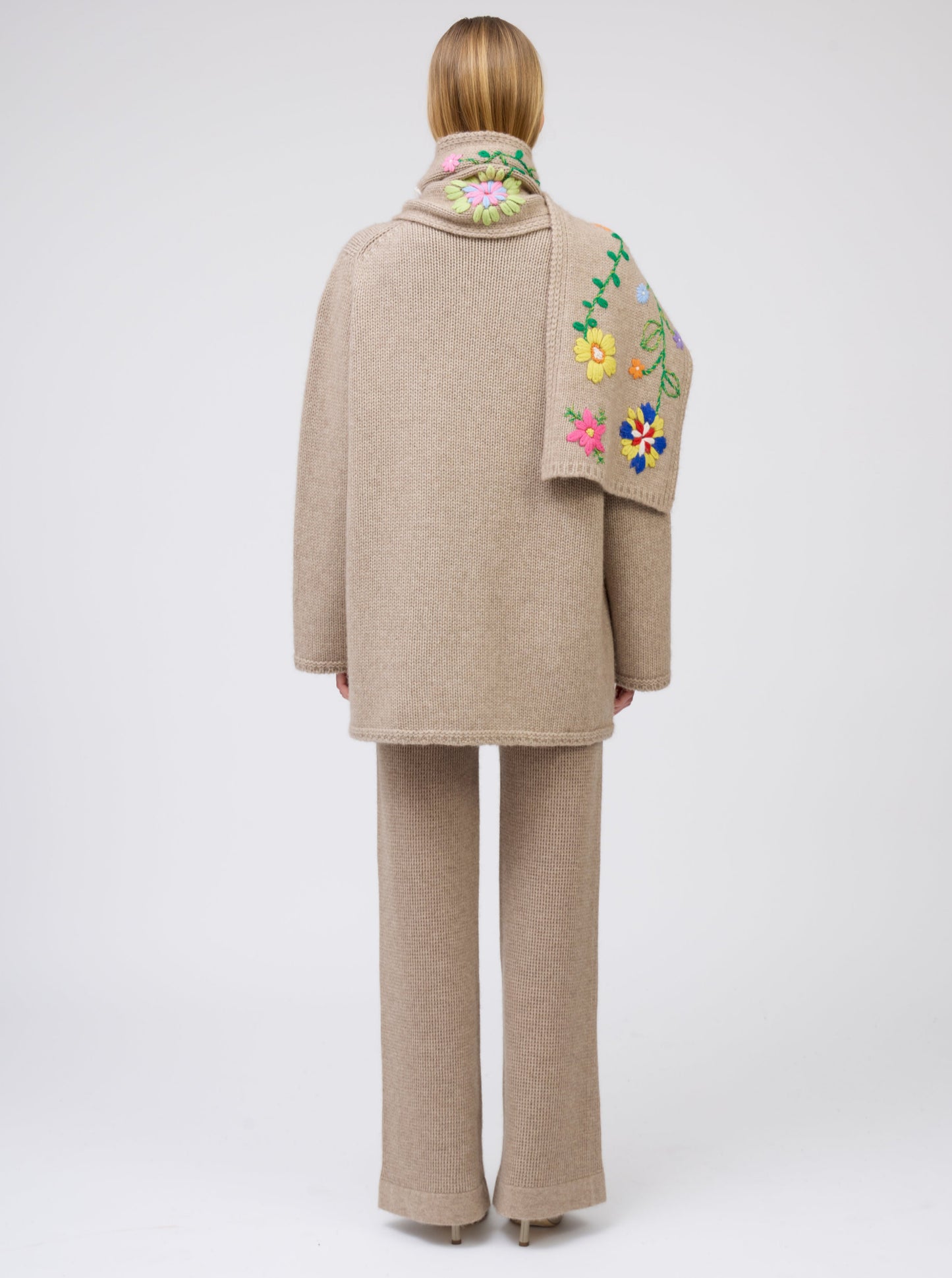 Back view model wearing light brown cashmere hip-length jacket with brightly coloured hand-embroidered scarf over one shoulder; worn with matching light brown knit pants.