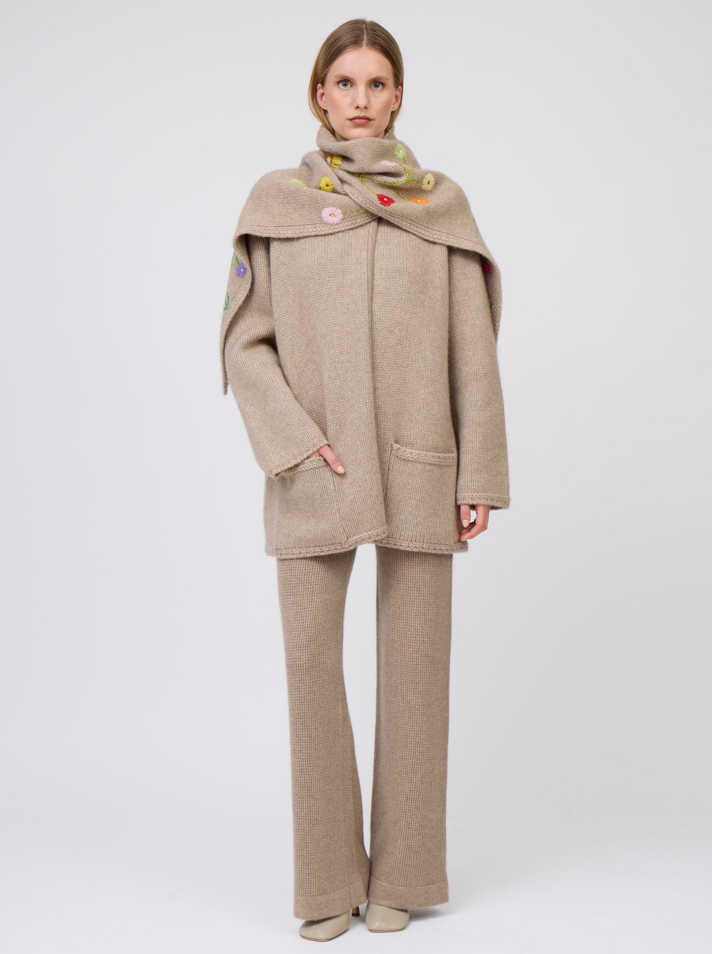 Front view model wearing light brown cashmere hip-length jacket with brightly coloured hand-embroidered scarf wrapped over shoulders; with one hand in pocket and worn with matching light brown knit pants.