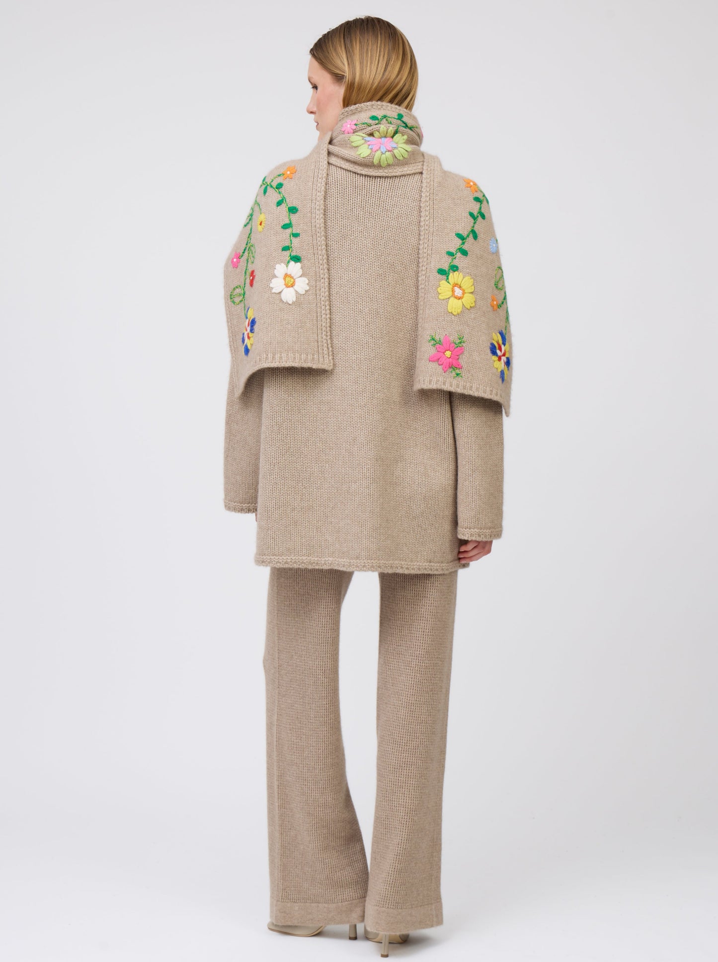 Back view model wearing light brown cashmere hip-length jacket with brightly coloured hand-embroidered floral scarf thrown over both shoulders; worn with matching light brown knit pants.