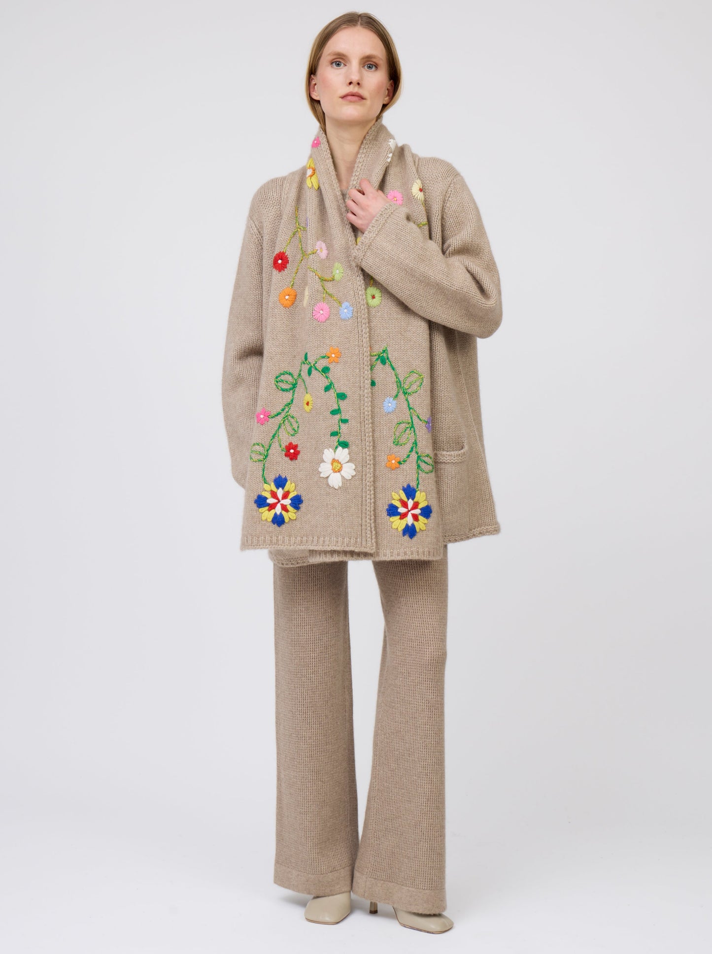 Front view model wearing light brown cashmere hip-length jacket with brightly coloured hand-embroidered scarf worn down the centre front of jacket; worn with matching light brown knit pants.