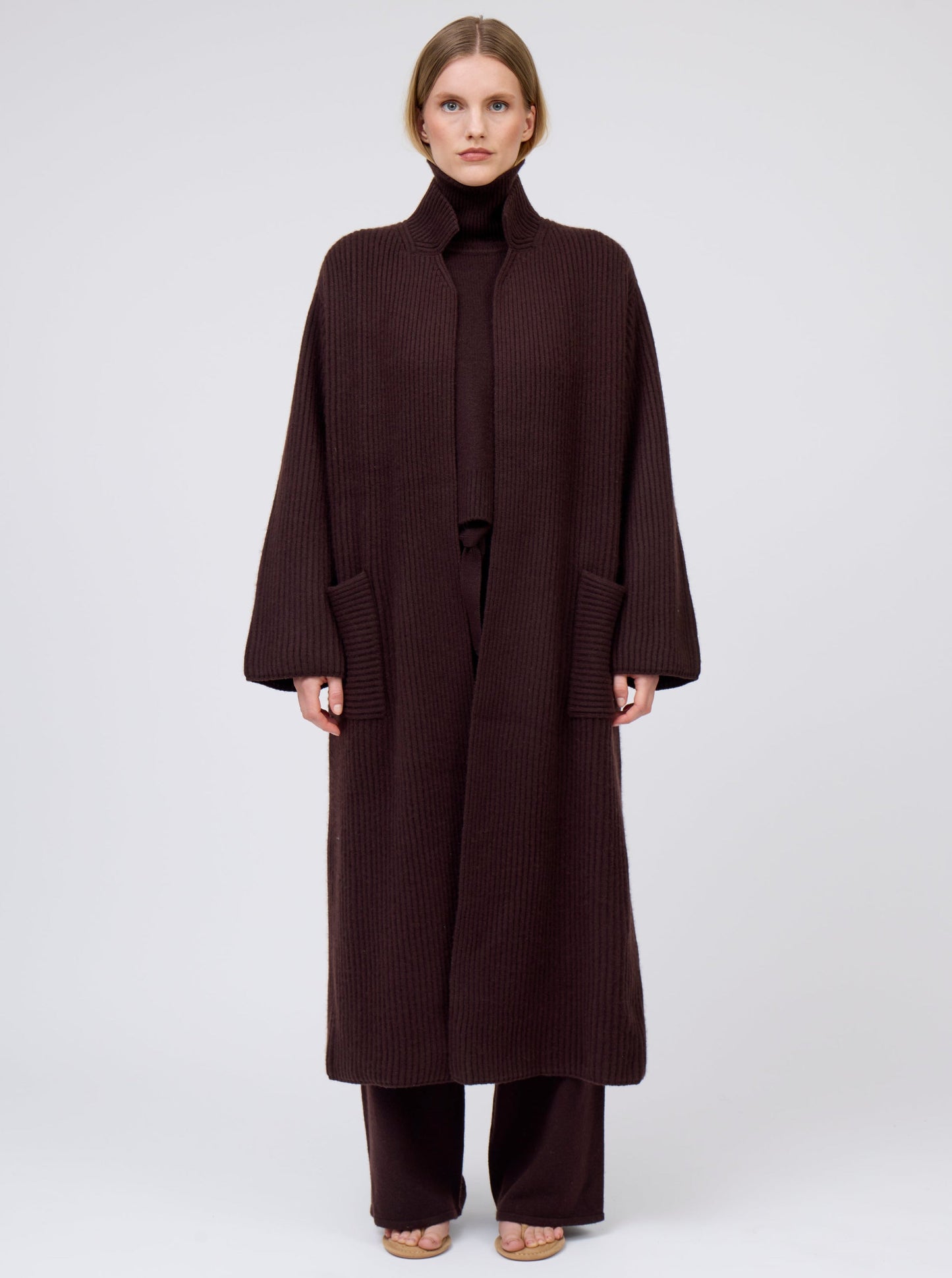 Model wearing a long cardigan coat in dark brown ribbed knit cashmere: straight cut with a relaxed fit, deep patch pockets. Worn with dark mahogany brown roll neck oversize jumper and wide leg pants.