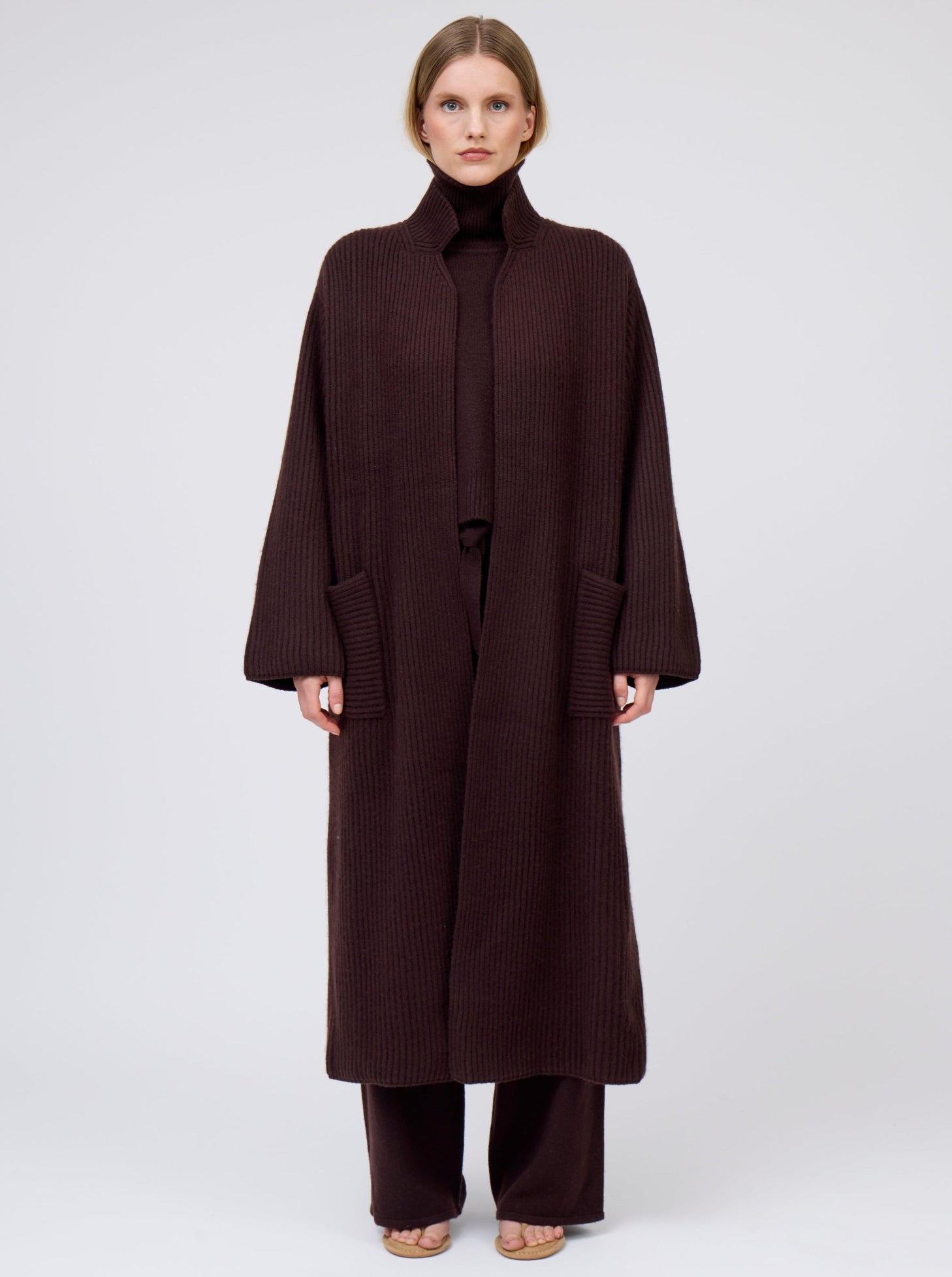 Model wearing a long cardigan coat in dark brown  ribbed knit cashmere: straight cut with a relaxed fit, deep patch pockets. Worn with dark mahogany brown roll neck oversize jumper and wide leg pants.