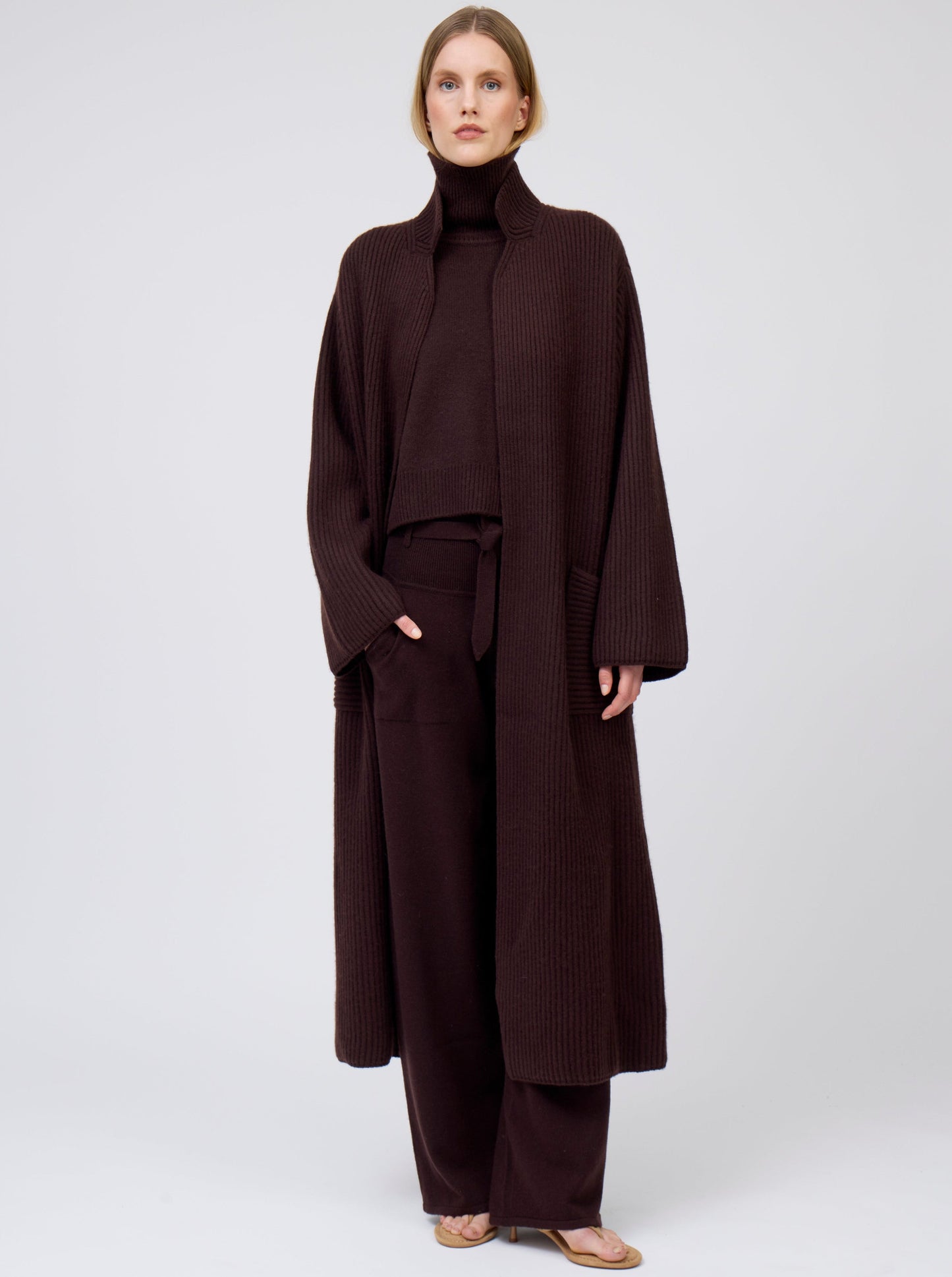 Model wearing a long cardigan coat in ribbed knit dark brown cashmere. Straight cut with a relaxed fit, deep patch pockets; standing with her hand in one pocket of matching dark brown cashmere pants. Worn with dark mahogany brown roll neck oversize jumper.