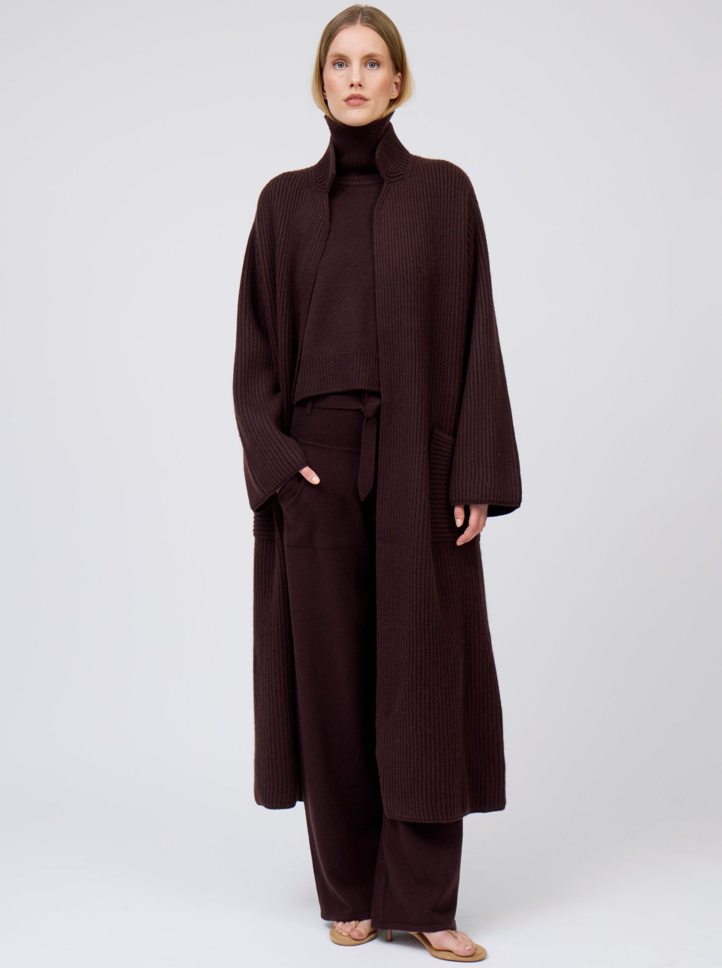 Model wearing a long cardigan coat in ribbed knit dark brown cashmere. Straight cut with a relaxed fit, deep patch pockets; standing with her hand in one pocket of matching dark brown cashmere pants. Worn with dark mahogany brown roll neck oversize jumper.