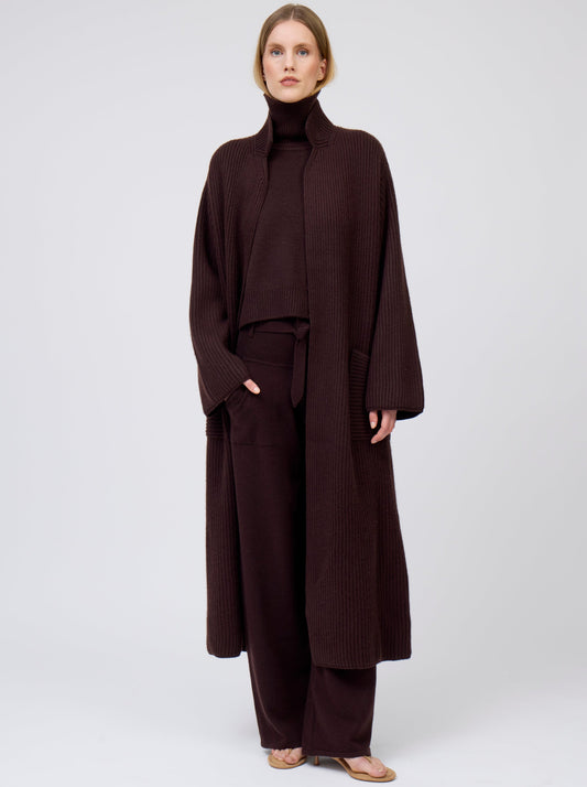 Model wearing a long cardigan coat in ribbed knit dark brown cashmere. Straight cut with a relaxed fit, deep patch pockets; standing with her hand in one pocket of matching dark brown cashmere pants. Worn with dark mahogany brown roll neck oversize jumper.