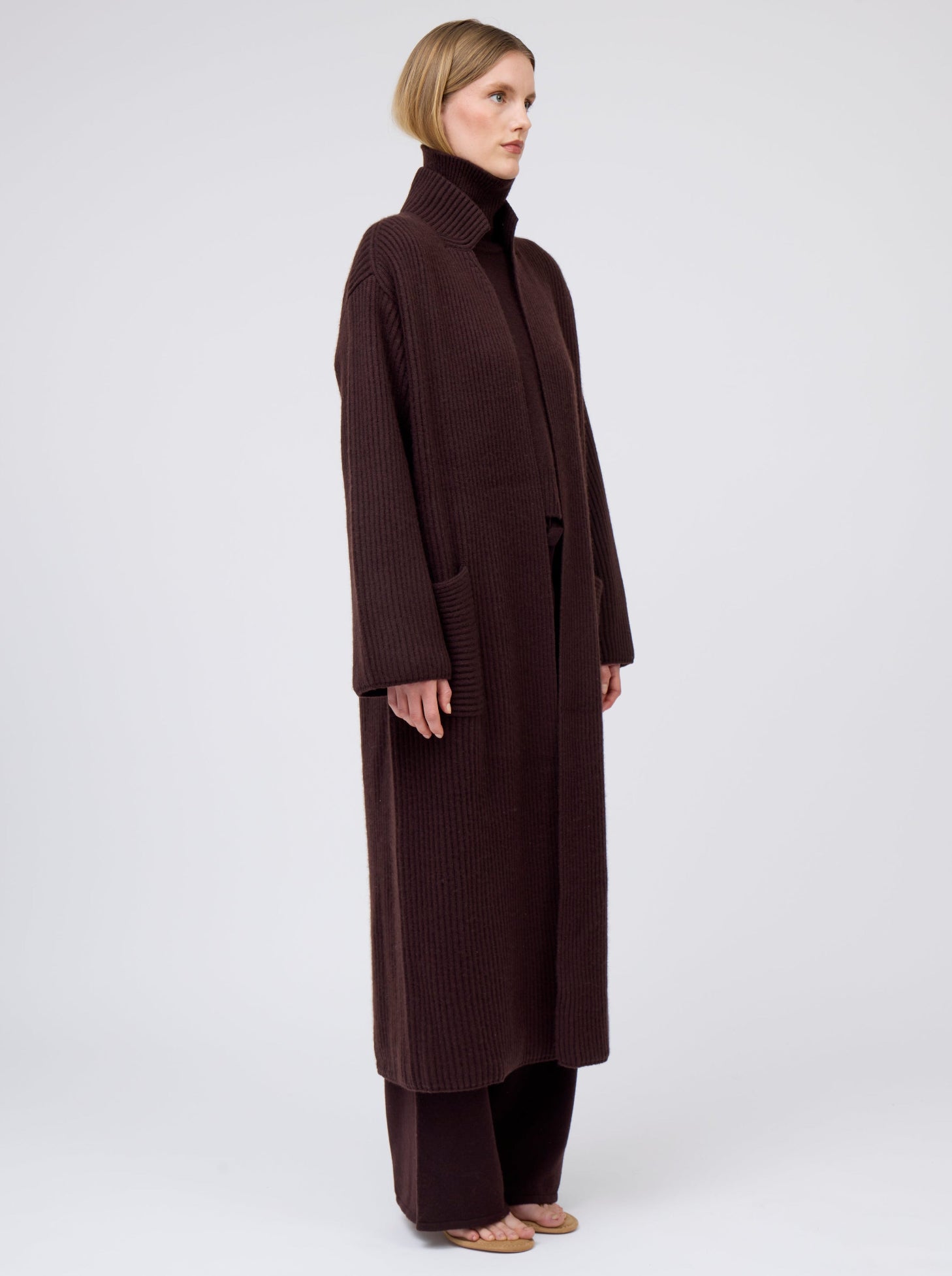 Side view of model wearing a long cardigan coat in dark brown  ribbed knit cashmere: with a relaxed fit and patch pockets. Worn with dark mahogany brown roll neck oversize jumper and wide leg pants.