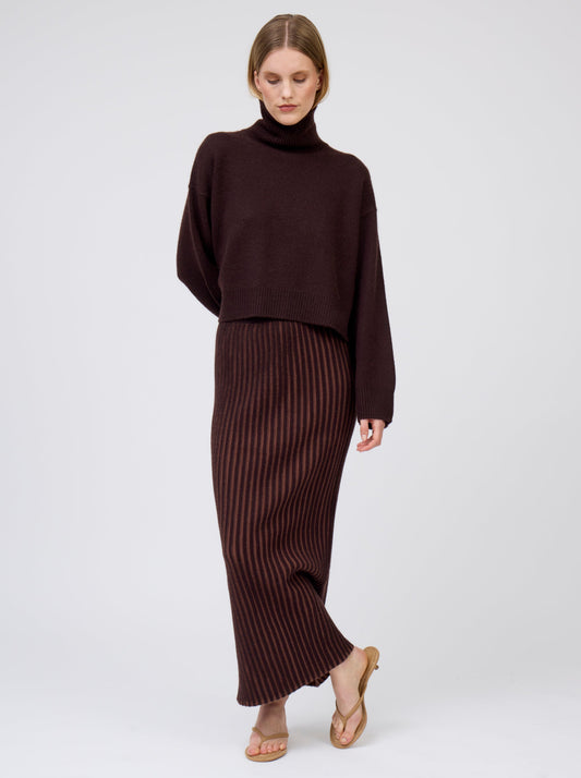 Front view of model wearing slim cashmere jersey maxi skirt in vertical stripes of mahogany and chocolate brown. Worn with cropped length mahogany brown roll neck jumper.
