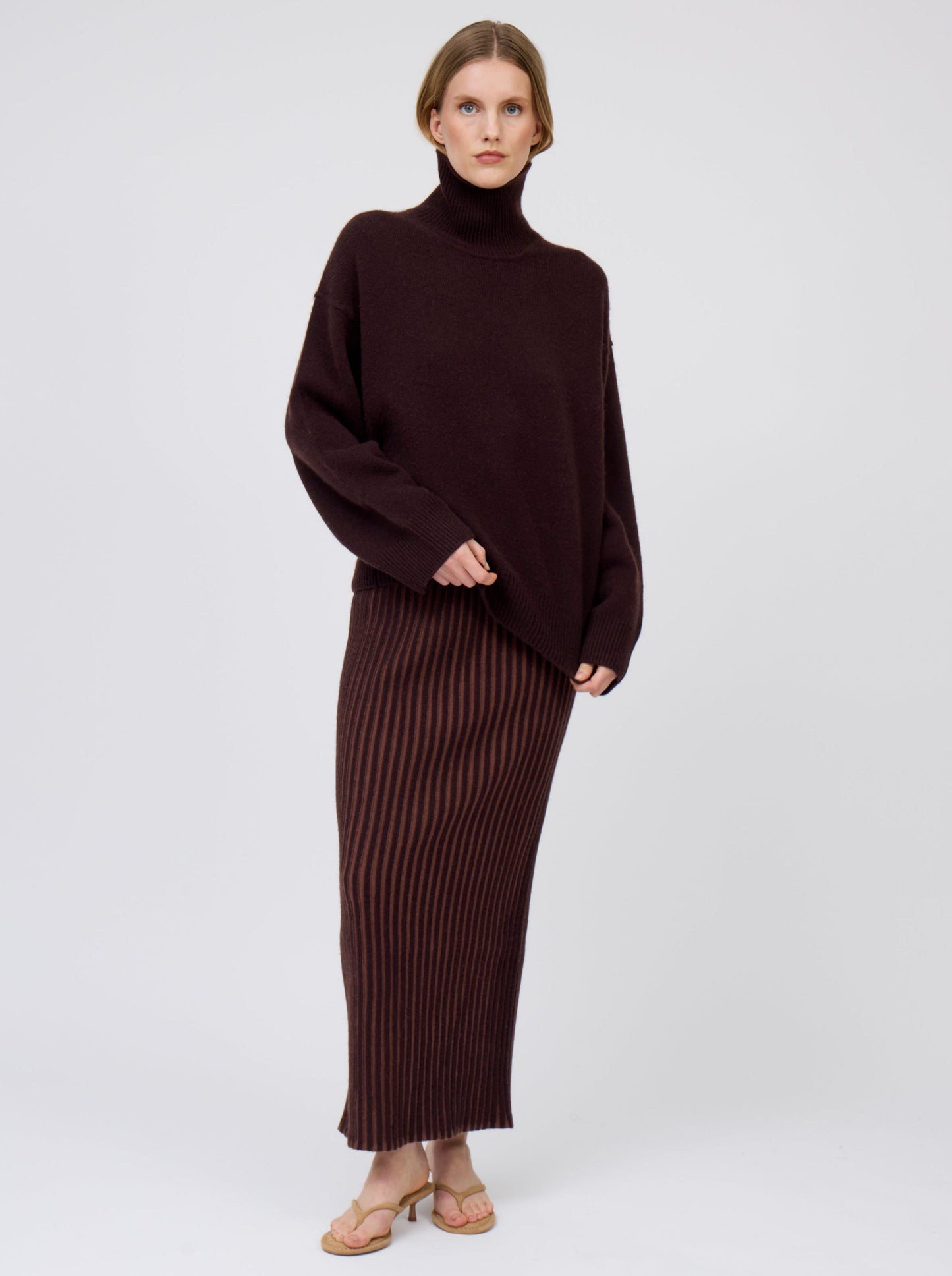 Model wears slim cashmere jersey maxi skirt in vertical stripes of mahogany and chocolate brown. Worn with oversized mahogany brown roll neck jumper.