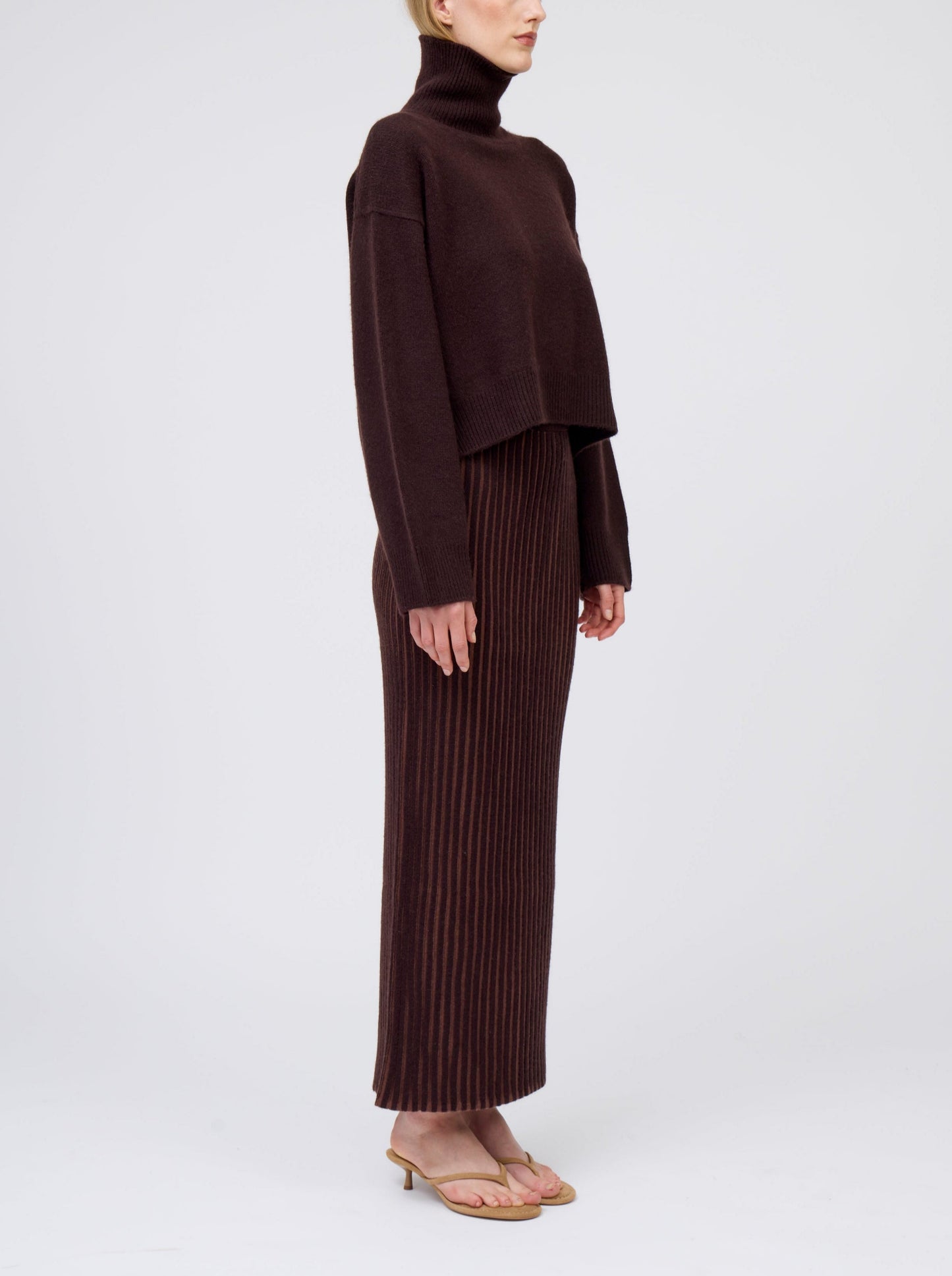 Side view of model wearing slim cashmere jersey maxi skirt in vertical stripes of mahogany and chocolate brown. Worn with cropped length mahogany brown roll neck jumper.