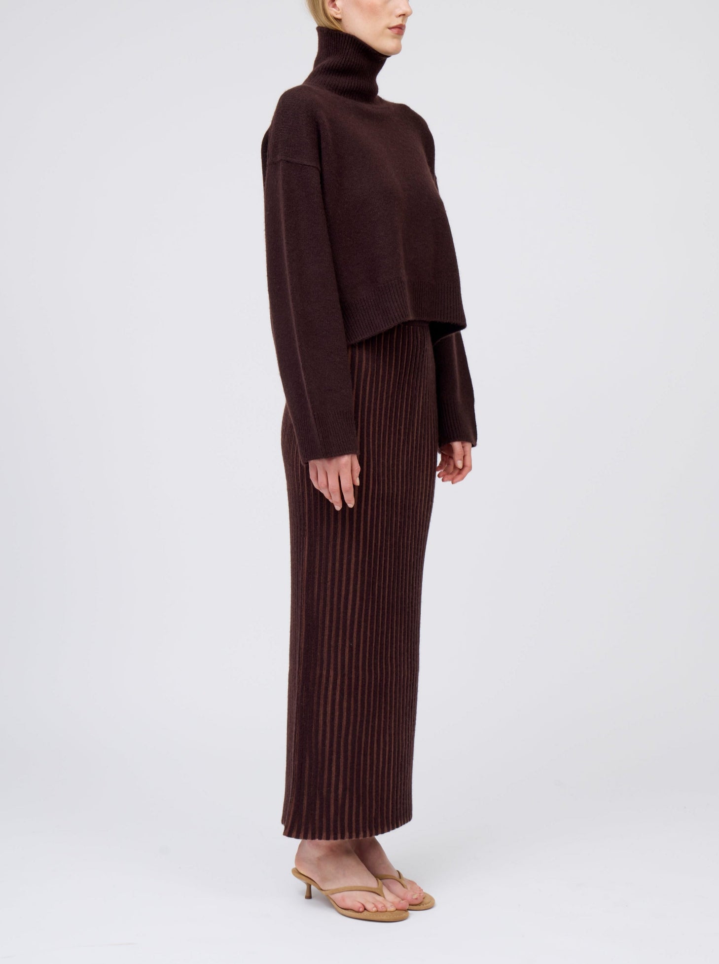 Side view of model wearing slim cashmere jersey maxi skirt in vertical stripes of mahogany and chocolate brown. Worn with cropped length mahogany brown roll neck jumper.