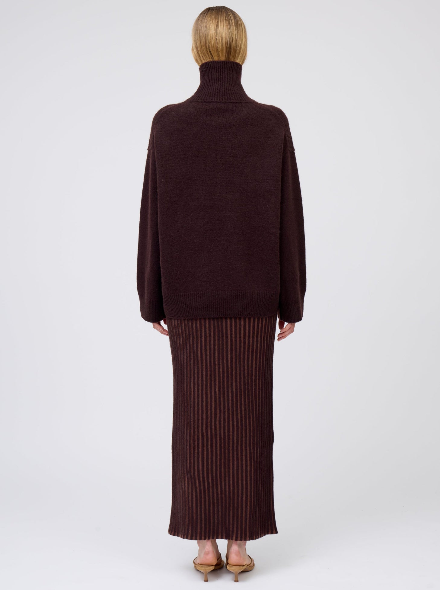 Back view of model wearing slim cashmere jersey maxi skirt in vertical stripes of mahogany and chocolate brown. Worn with oversized mahogany brown roll neck jumper.