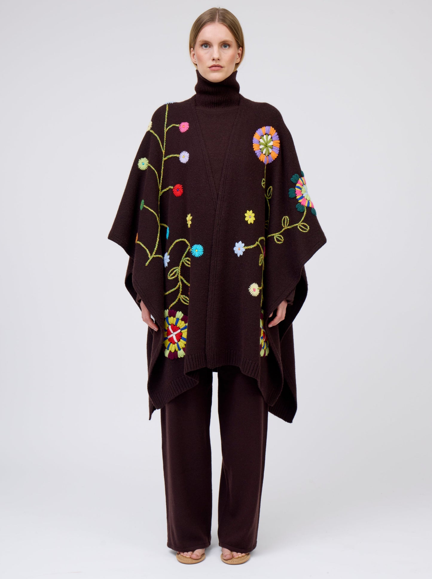 Front view model wearing mahogany brown cashmere mid-length wrap with brightly coloured hand-embroidery; worn with matching brown jumper and brown pants.
