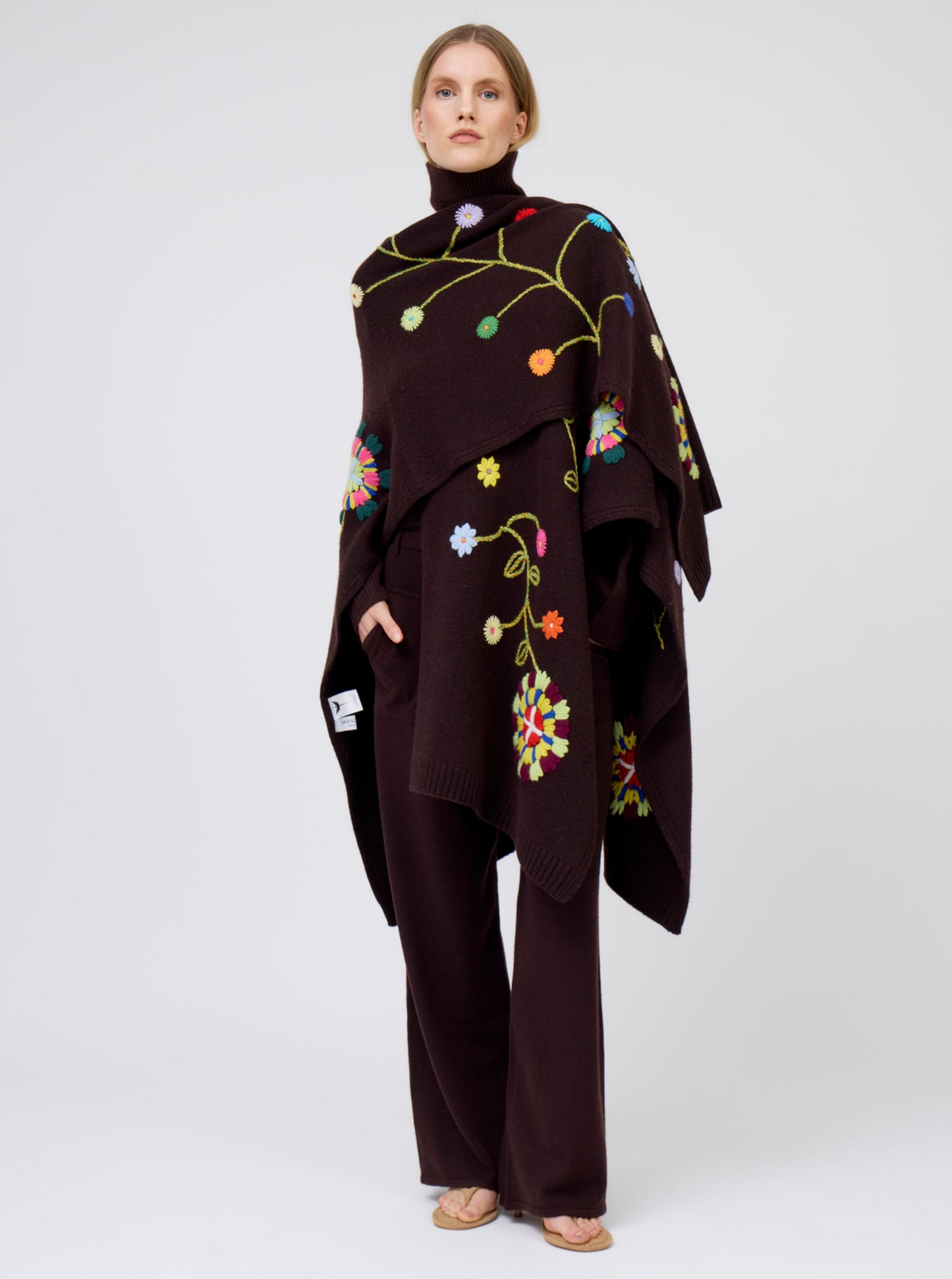 Model wears mahogany brown cashmere mid-length wrap with brightly coloured floral hand-embroidery, with one side thrown over shoulder; worn with matching brown jumper and brown pants.