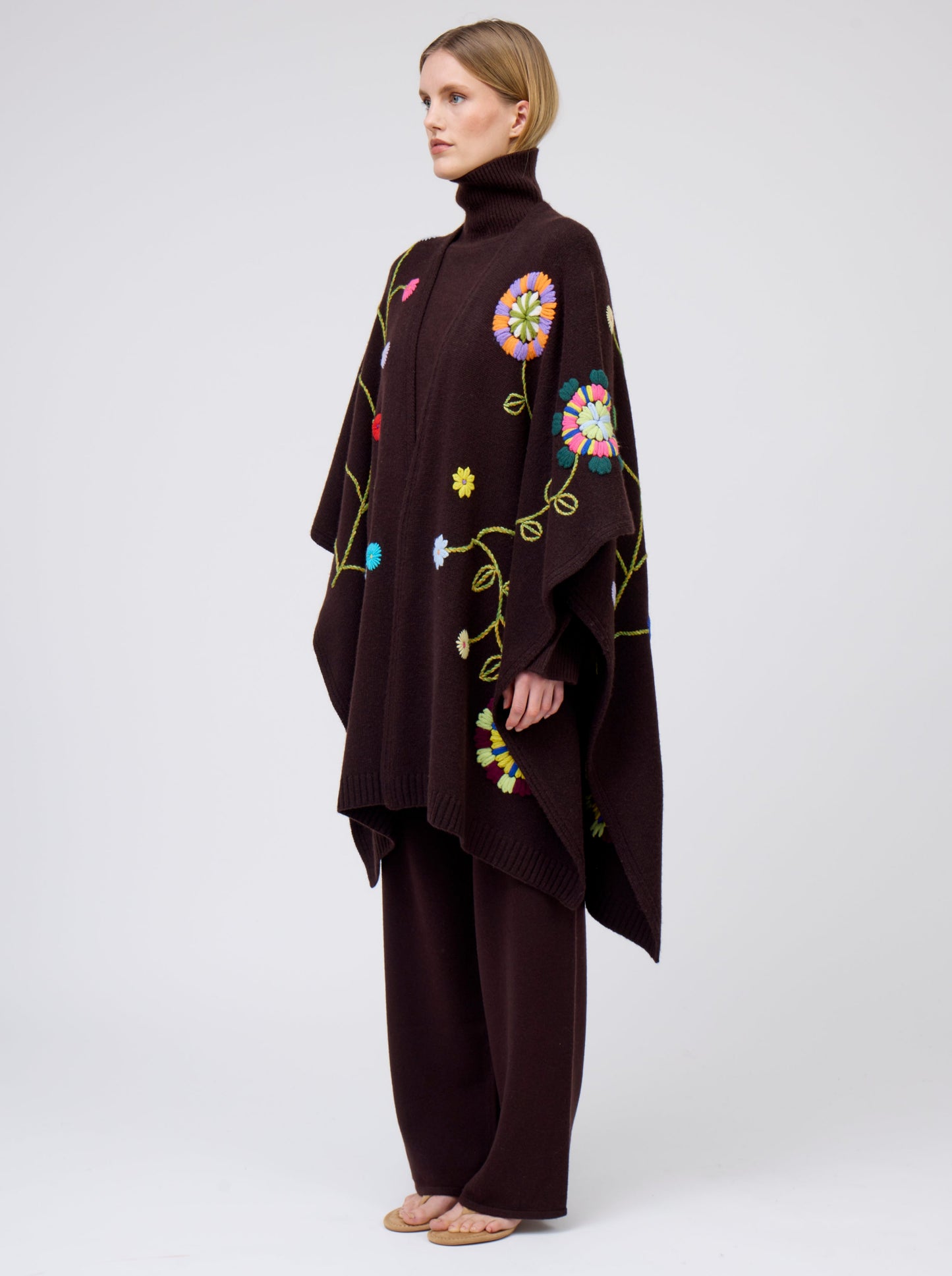 Side view model wearing mahogany brown cashmere mid-length wrap with brightly coloured floral hand-embroidery; worn with matching brown jumper and brown pants.