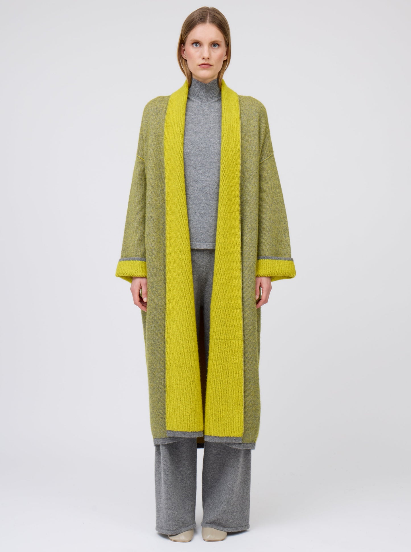 Front straight view model in long mustard yellow and grey mix cashmere knit coat with yellow collar revere and turn back cuffs; wearing with grey knit top and wide leg cashmere pants.